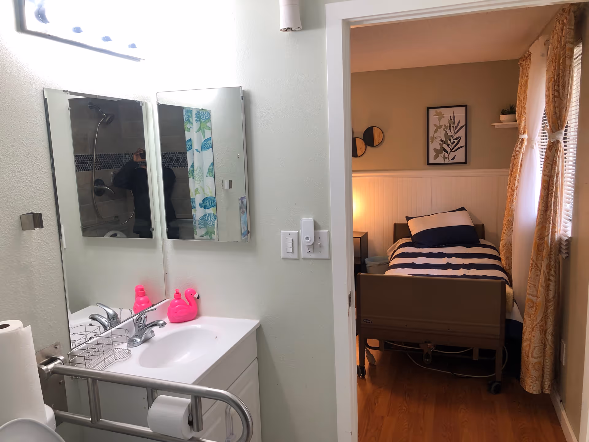 Bathroom sink and mirror with a grab bar and pink soap dispensers, view through a doorway into a small bedroom with a single bed.