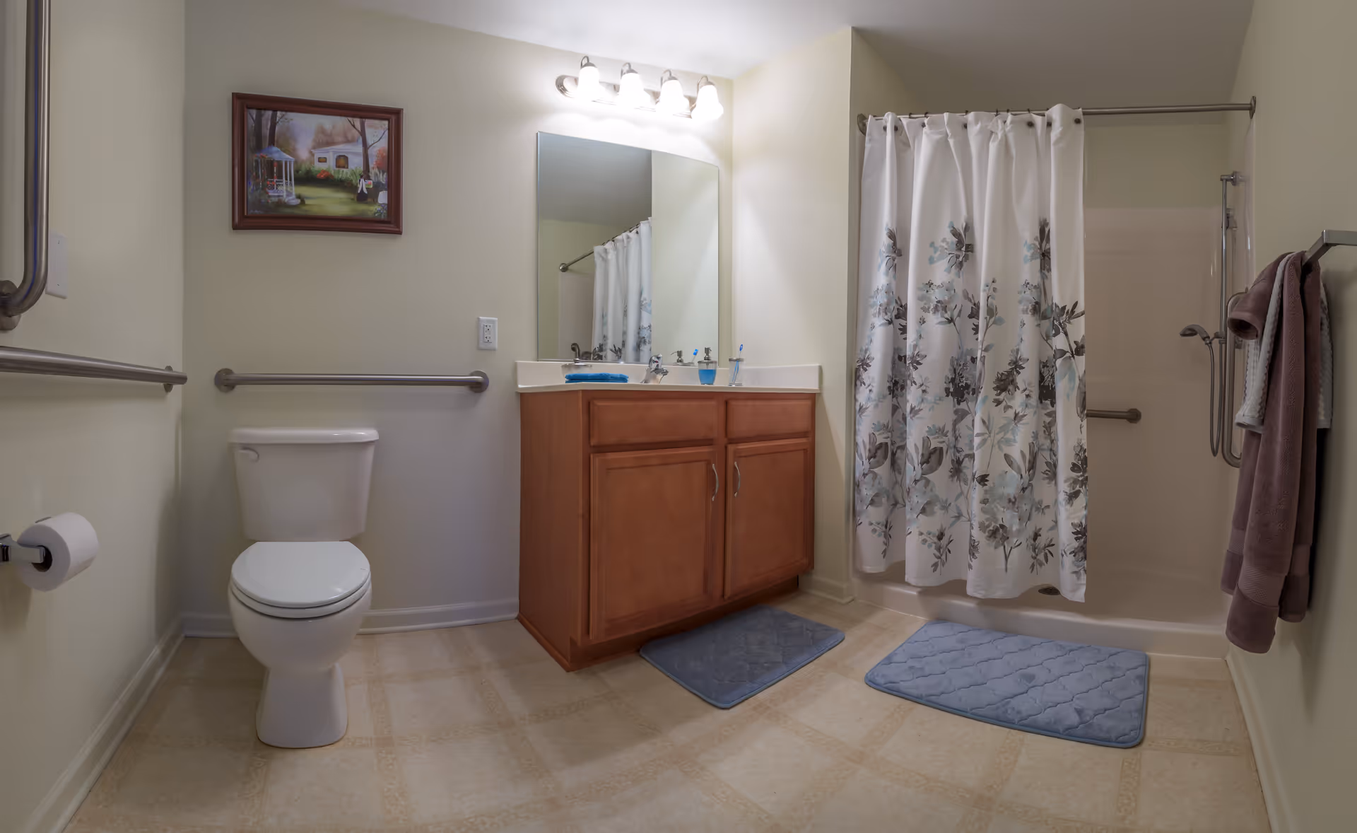 A clean and accessible bathroom featuring a toilet with grab bars on the walls, a wooden vanity with a large mirror and overhead lights, a shower with a floral-patterned curtain, two blue bath mats on the floor, and a towel rack holding folded towels. A framed painting hangs on the wall above the toilet.