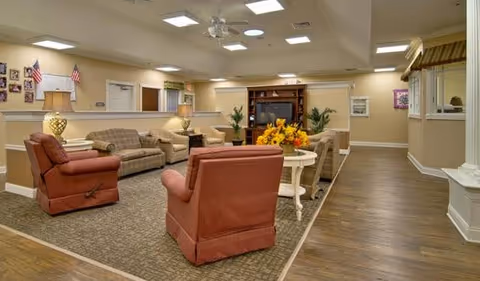 A spacious communal lounge with sofas, armchairs, a television cabinet, and coffee tables in a senior living facility.