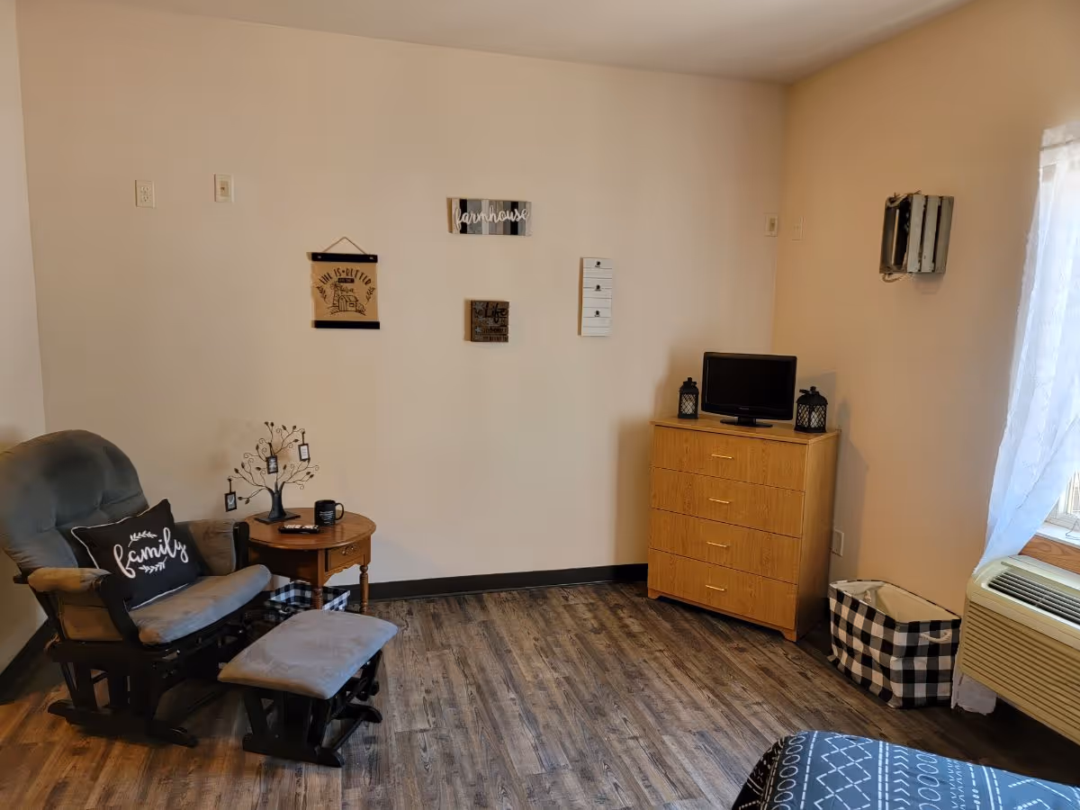A cozy living area with a cushioned rocking chair and matching footrest, a small wooden side table with decorative items, a wooden dresser with a small TV on top, and a window with sheer white curtains. The walls are decorated with small framed signs, and the floor has wood-like vinyl flooring.