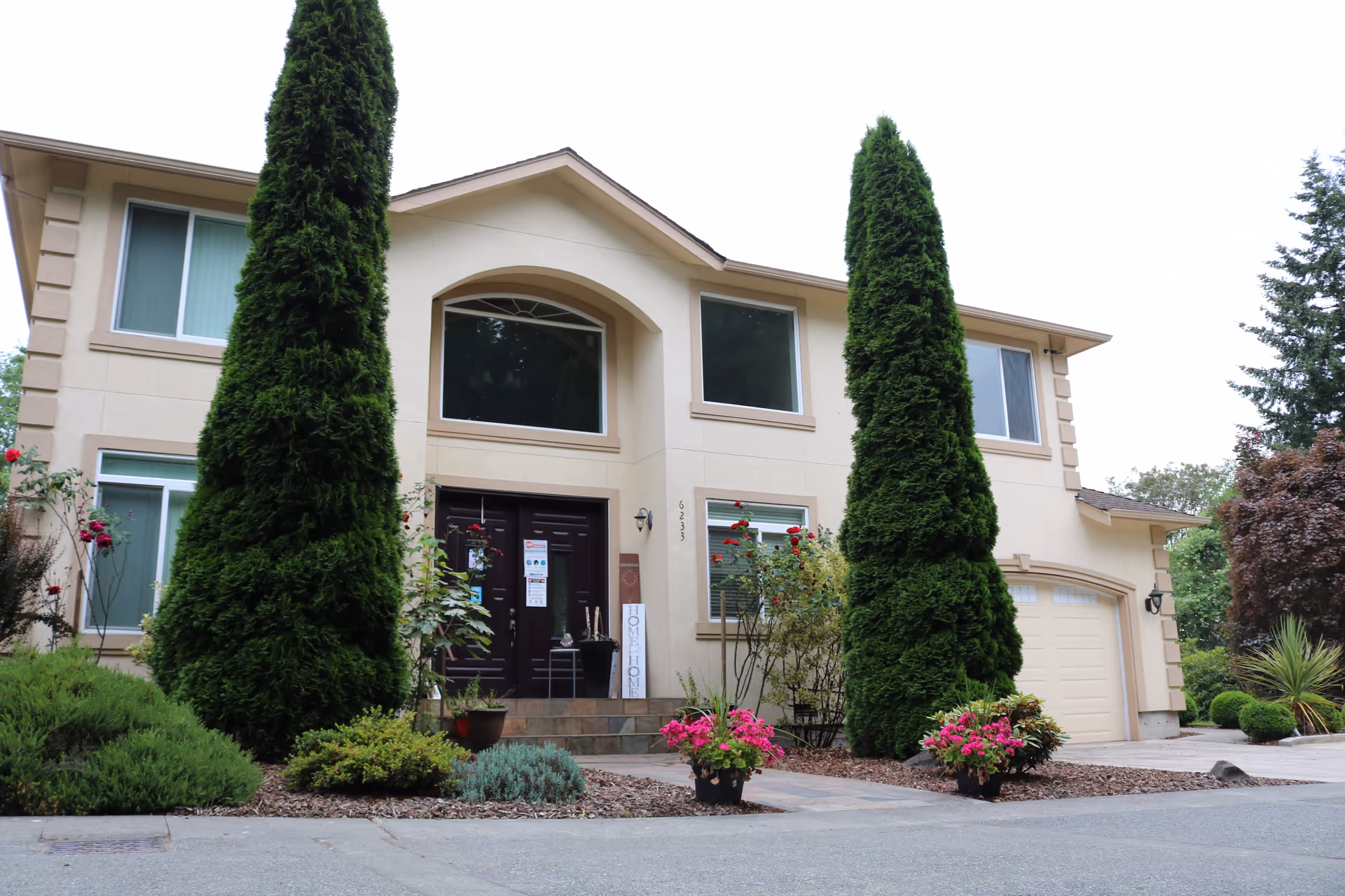 Front exterior view of a two-story beige house with a dark brown double front door, large windows, and a garage on the right side. The entrance is flanked by two tall, narrow evergreen trees and surrounded by various shrubs and flowering plants in pots and garden beds.