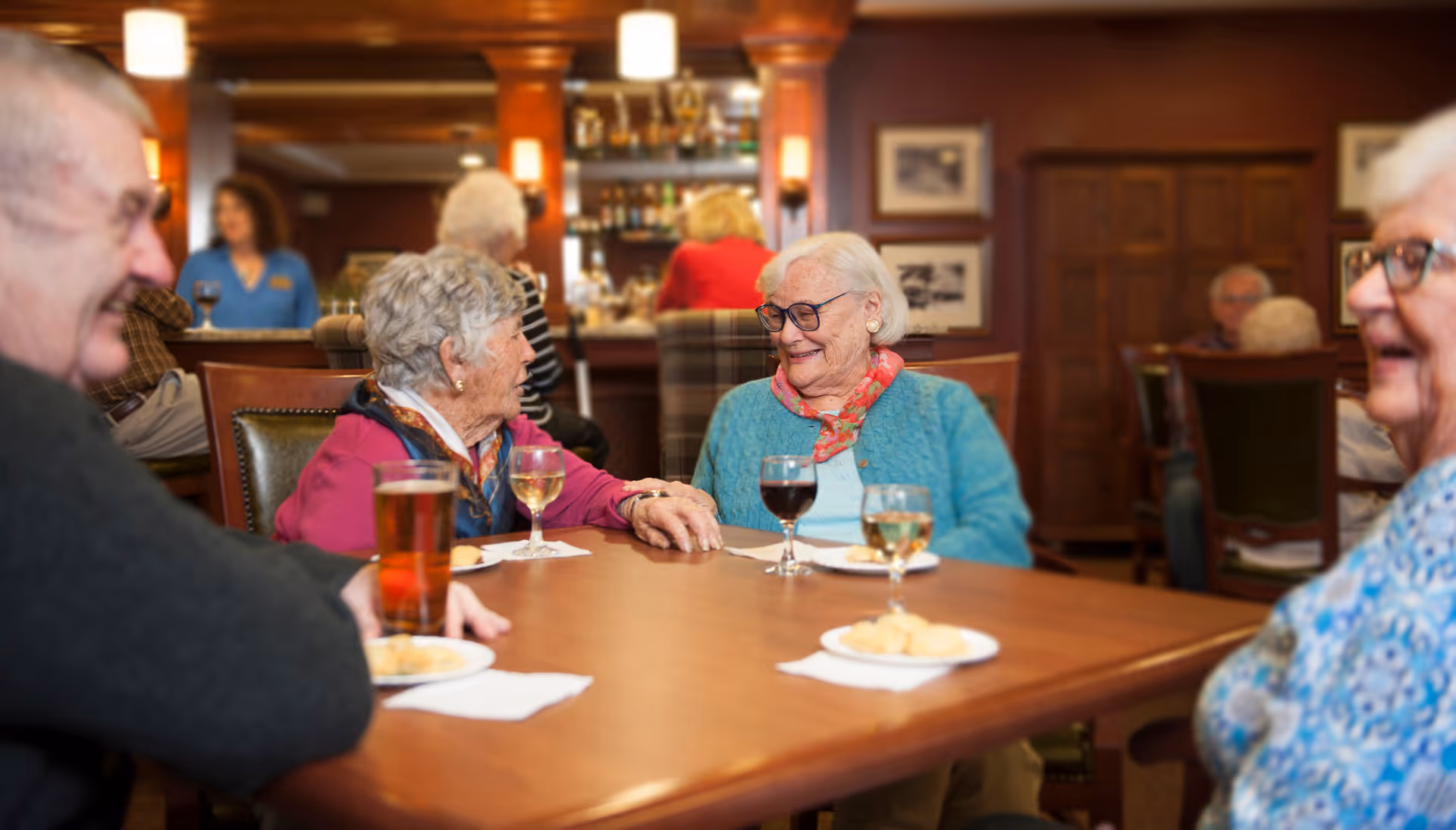 A group of elderly people sitting around a wooden table in a cozy dining or lounge area, enjoying drinks and snacks while engaging in conversation. The background shows a bar area with bottles and glasses, warm lighting, and wooden decor.