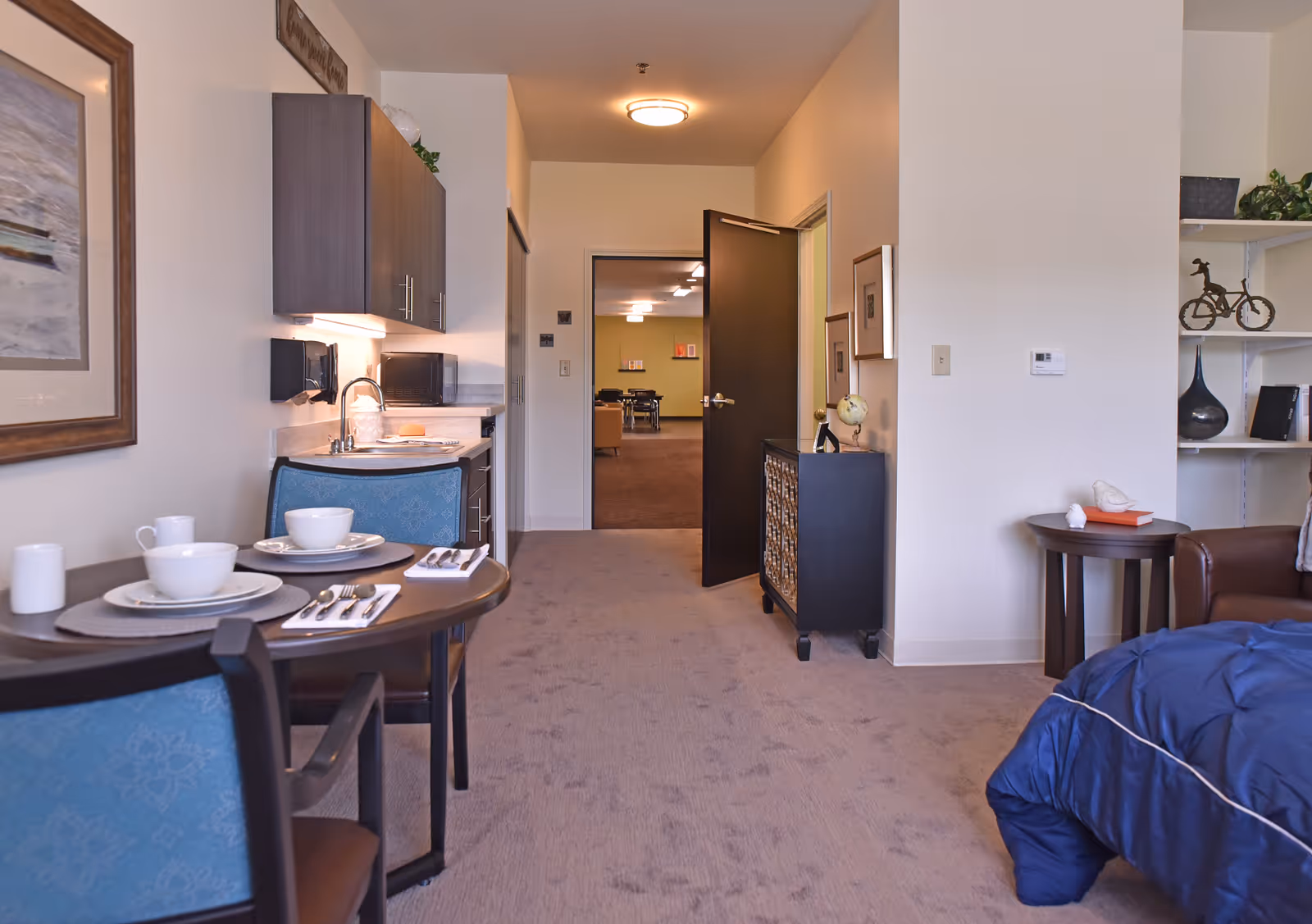 Interior view of a senior living facility room at The Glen showing a small dining table set for two with dishes and utensils, a kitchenette with cabinets and a sink, a partially visible bed with blue bedding, a side table with decorative items, and an open door leading to another room.