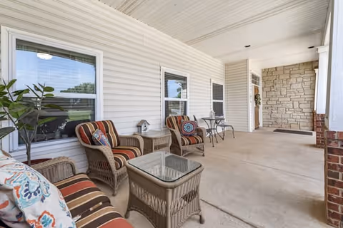 A spacious covered porch with beige siding and stone accents, furnished with wicker chairs and a glass-top wicker table. There are colorful cushions on the chairs and a small round metal table with two chairs further down the porch. A potted plant is placed near the window.