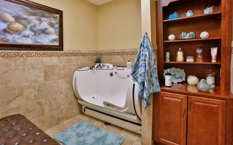 A bathroom featuring a walk-in bathtub with handrails and controls, beige tiled walls with a decorative border, a blue bath mat on the floor, a wooden cabinet with shelves holding various toiletries and decorative items, a blue and white striped towel hanging on the side of the tub, and a framed picture of rocks and flowing water on the wall.