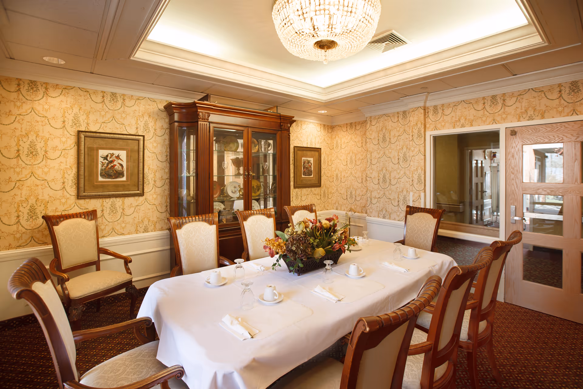 A formal dining room with a long table covered by a white tablecloth, set with cups, glasses, and napkins. The room features upholstered wooden chairs around the table, a chandelier hanging from the ceiling, patterned wallpaper, framed artwork on the walls, and a wooden china cabinet displaying dishes.