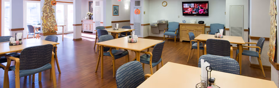 A bright and clean dining room in a senior living facility with several wooden tables and blue cushioned chairs arranged neatly. The room has wood flooring, large windows letting in natural light, and Halloween decorations including pumpkin faces on the walls. A flat-screen TV is mounted on the wall, and there are condiment holders on the tables.