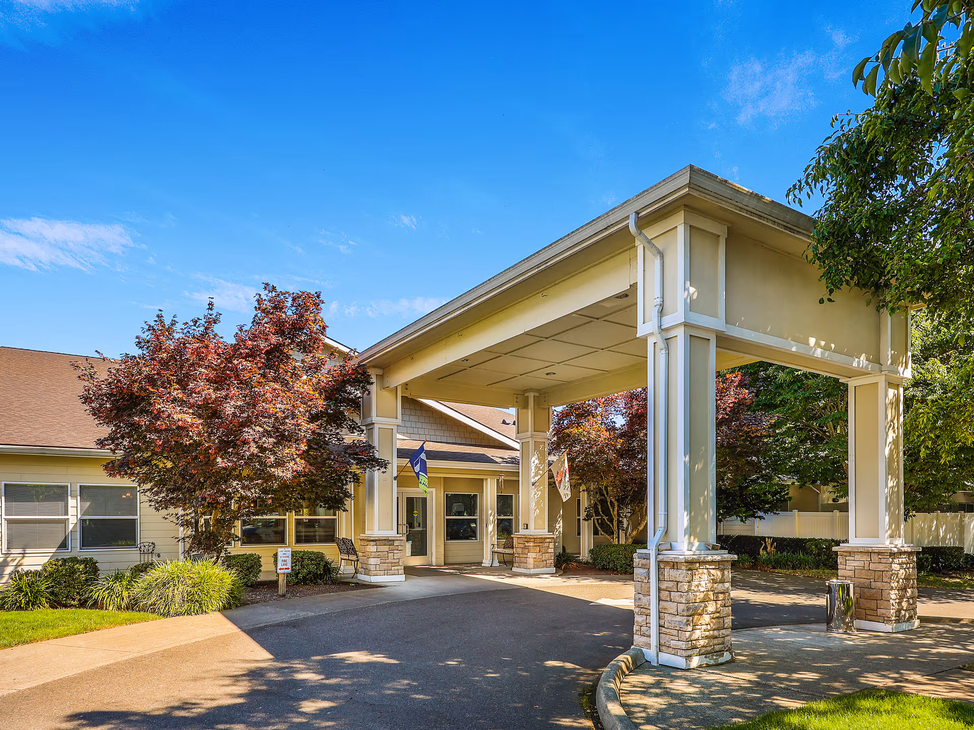 Entrance of Prestige Senior Living Auburn Meadows featuring a covered driveway supported by columns with stone bases, surrounded by trees and landscaping under a clear blue sky.