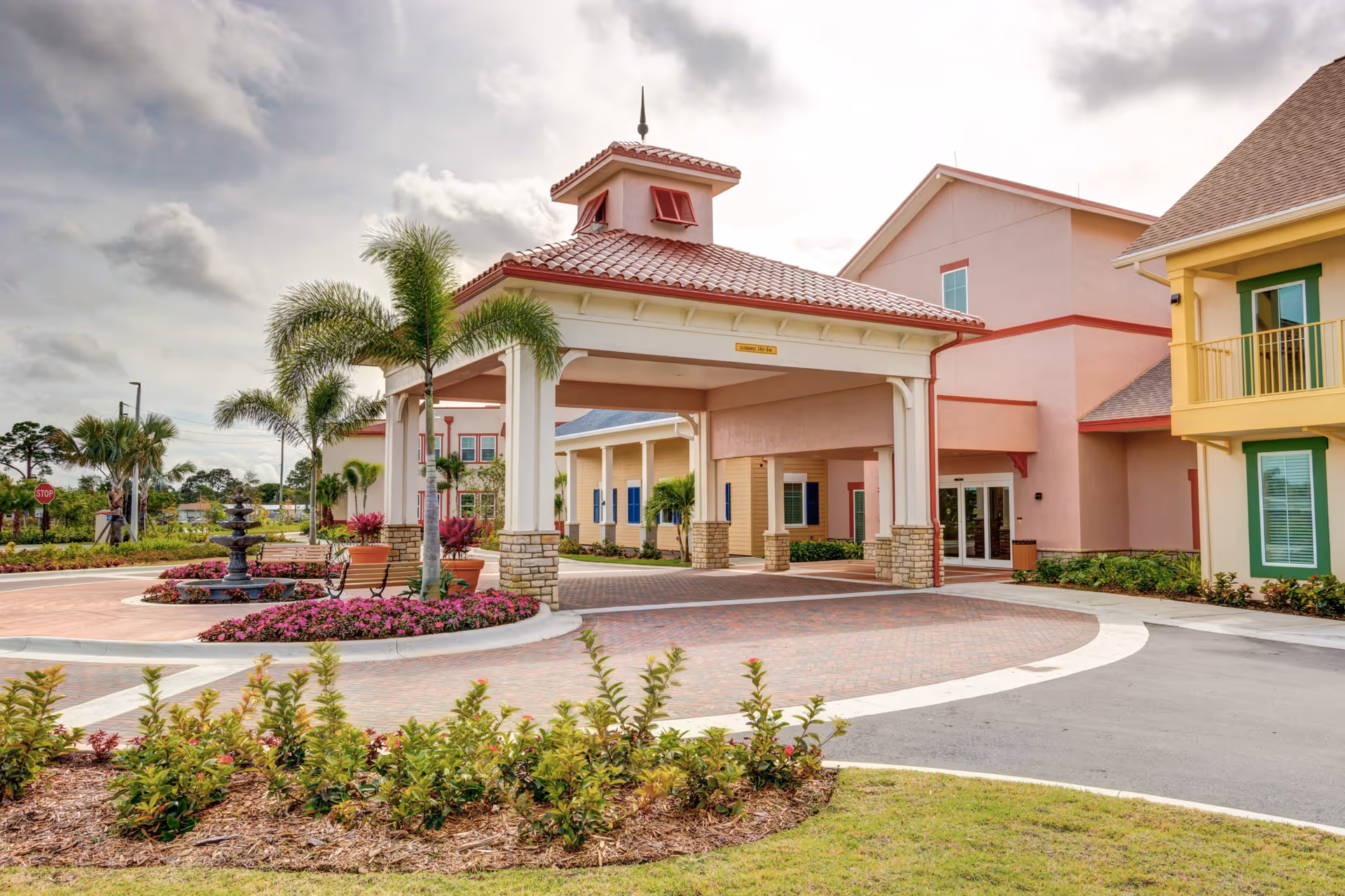 Covered porte-cochere entrance of a colorful senior living building with palm trees, a fountain, and a landscaped circular driveway.