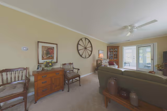 A cozy living room with beige walls and carpet. The room features a green sofa, two wooden chairs with cushions, a wooden sideboard with a flower arrangement and a phone, a large decorative wheel mounted on the wall, a framed painting, a floral armchair with a lamp beside it, a glass-front cabinet, and double glass doors leading to a sunroom or patio area.