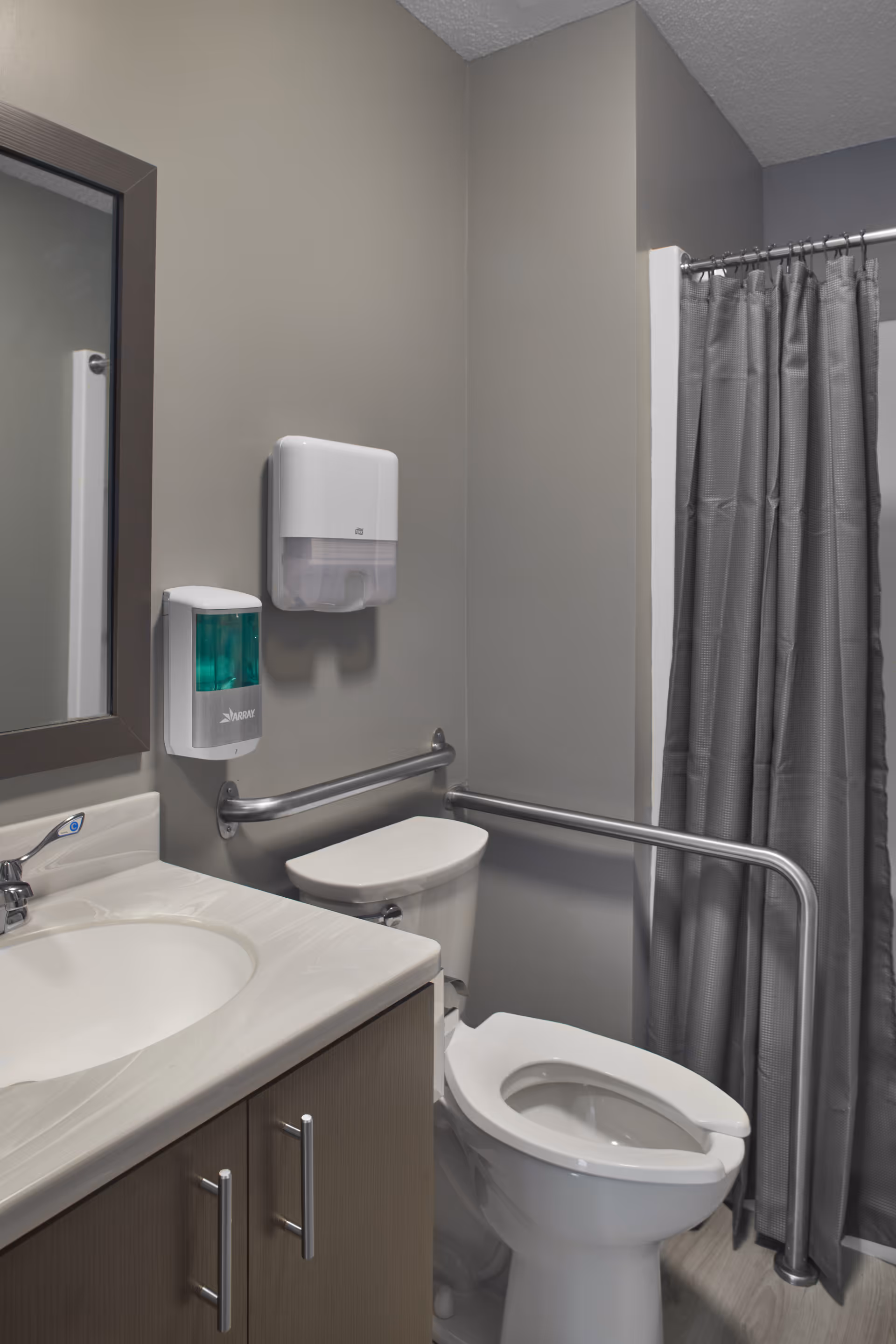 A bathroom with a white toilet, a sink with a countertop and cabinet, a mirror, a soap dispenser, a paper towel dispenser, and a shower area with a gray shower curtain. There are metal grab bars installed near the toilet for accessibility.