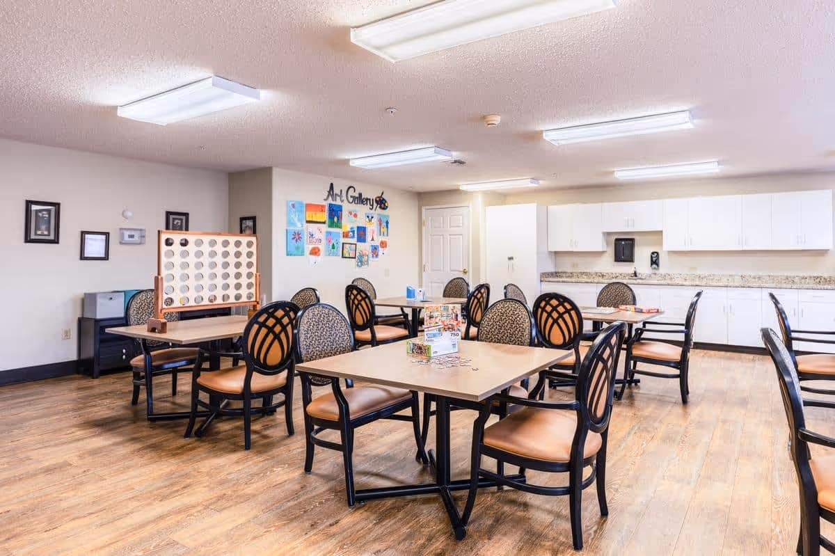 A bright and spacious activity room in a senior living facility with multiple tables and chairs arranged for group activities. The room features a wooden floor, white cabinets along one wall, and an art gallery display with colorful artwork on the far wall. A large Connect Four game is set up on one of the tables.
