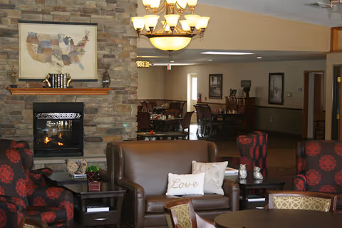 A cozy living room area with a stone fireplace, a brown leather loveseat with decorative pillows including one that says 'Love', two red and black patterned armchairs, and a round table with chairs. The room has warm lighting from a chandelier and there is a dining area visible in the background.