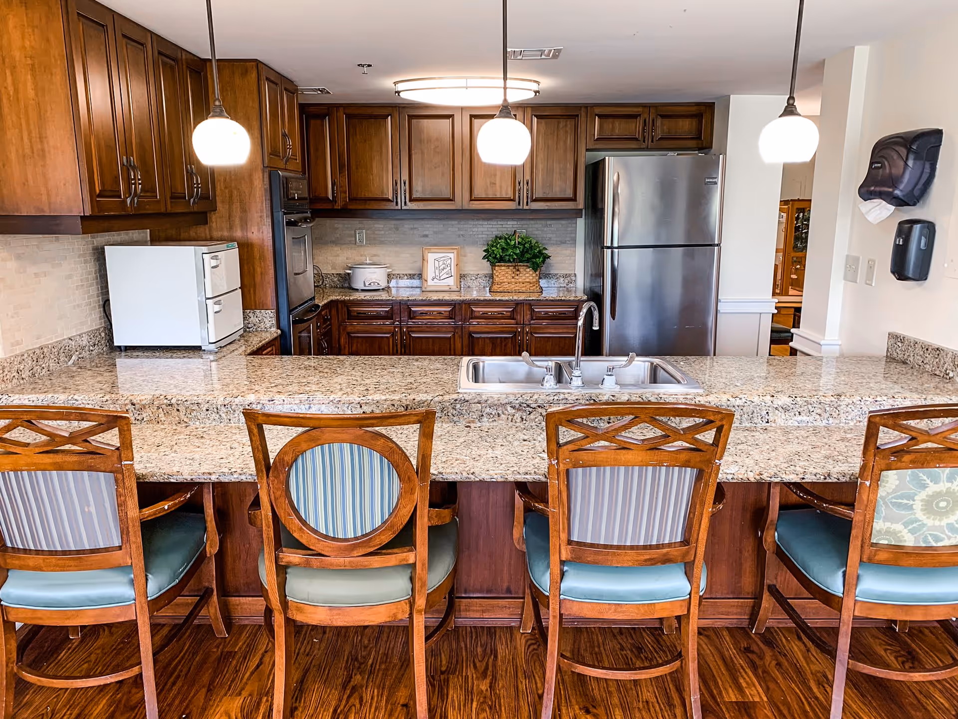 Granite-topped kitchen island with sink and four wooden bar stools in front of a wood-cabinet kitchen with stainless refrigerator and pendant lights.
