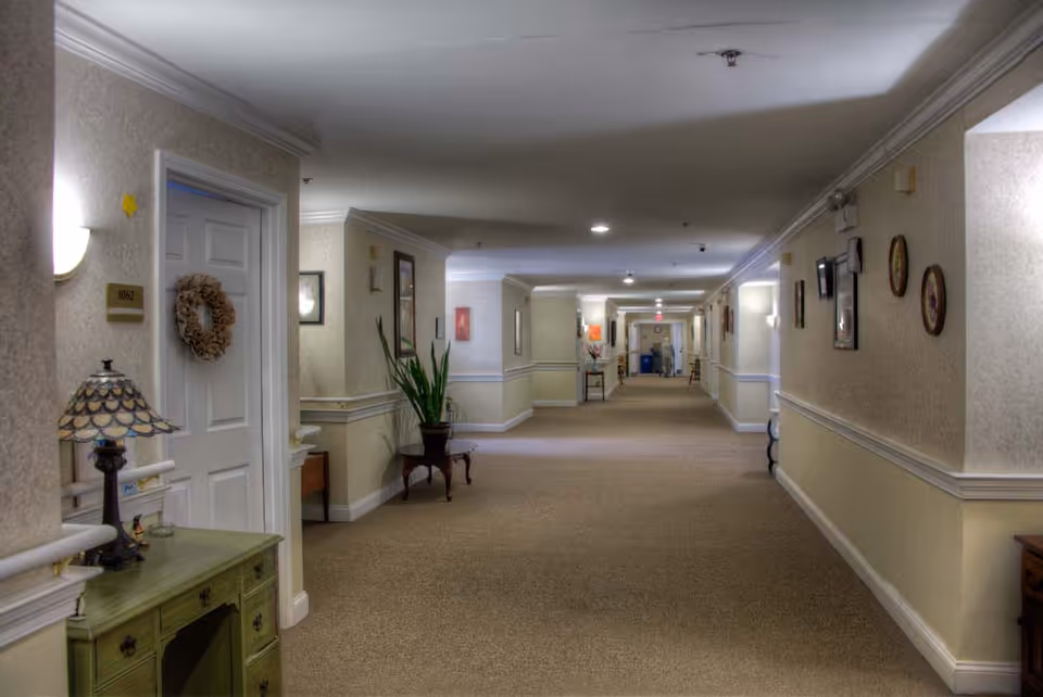 A long, carpeted hallway in an assisted living facility with beige walls and white trim. The hallway is decorated with framed pictures, a potted plant on a small table, and a lamp on a green cabinet near a door labeled 1062 with a wreath hanging on it. The corridor is well-lit with ceiling lights and wall sconces.