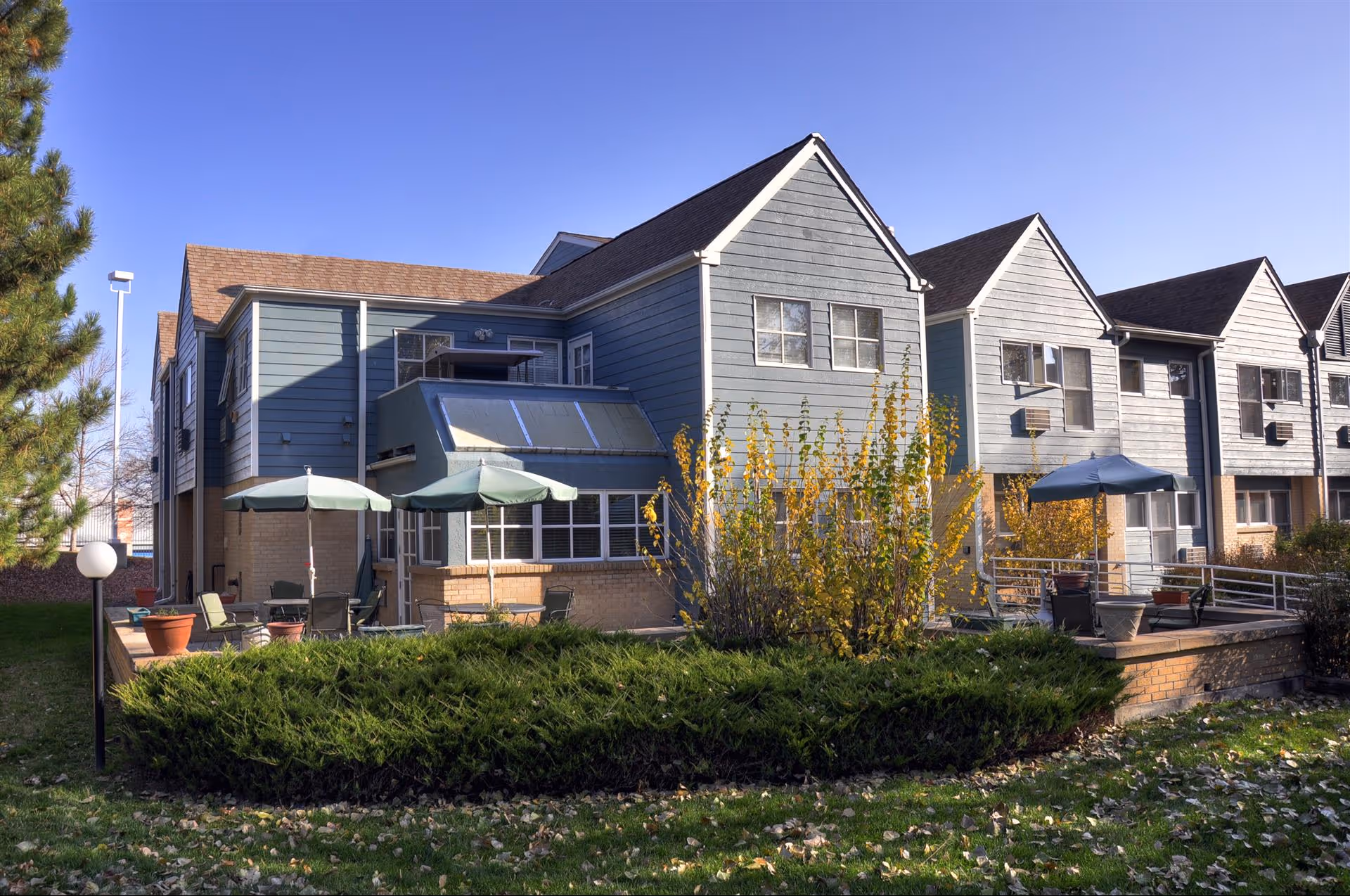 Exterior view of a multi-unit residential building with blue-gray siding and brown roofs under a clear blue sky. The building has a patio area with green umbrellas, outdoor chairs, and potted plants. There is greenery and bushes in the foreground and a tree on the left side.
