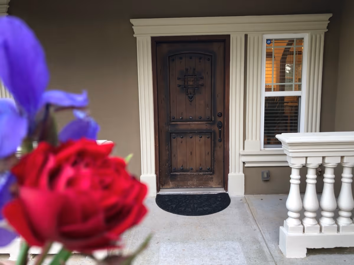 Front entrance of a building with a rustic wooden door framed by white decorative trim, a window with blinds to the right, and a white balustrade on the porch. In the foreground, there are blurred red and purple flowers.