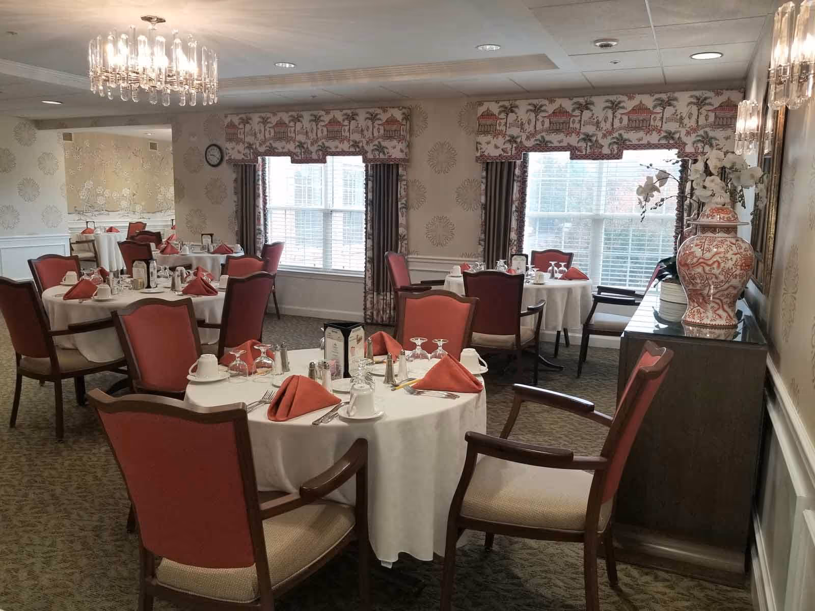 Dining room with round tables covered in white tablecloths, each set with red napkins, cups, glasses, and silverware. The room has red cushioned chairs, large windows with patterned valances, a chandelier, and a sideboard with a large decorative vase and flowers.