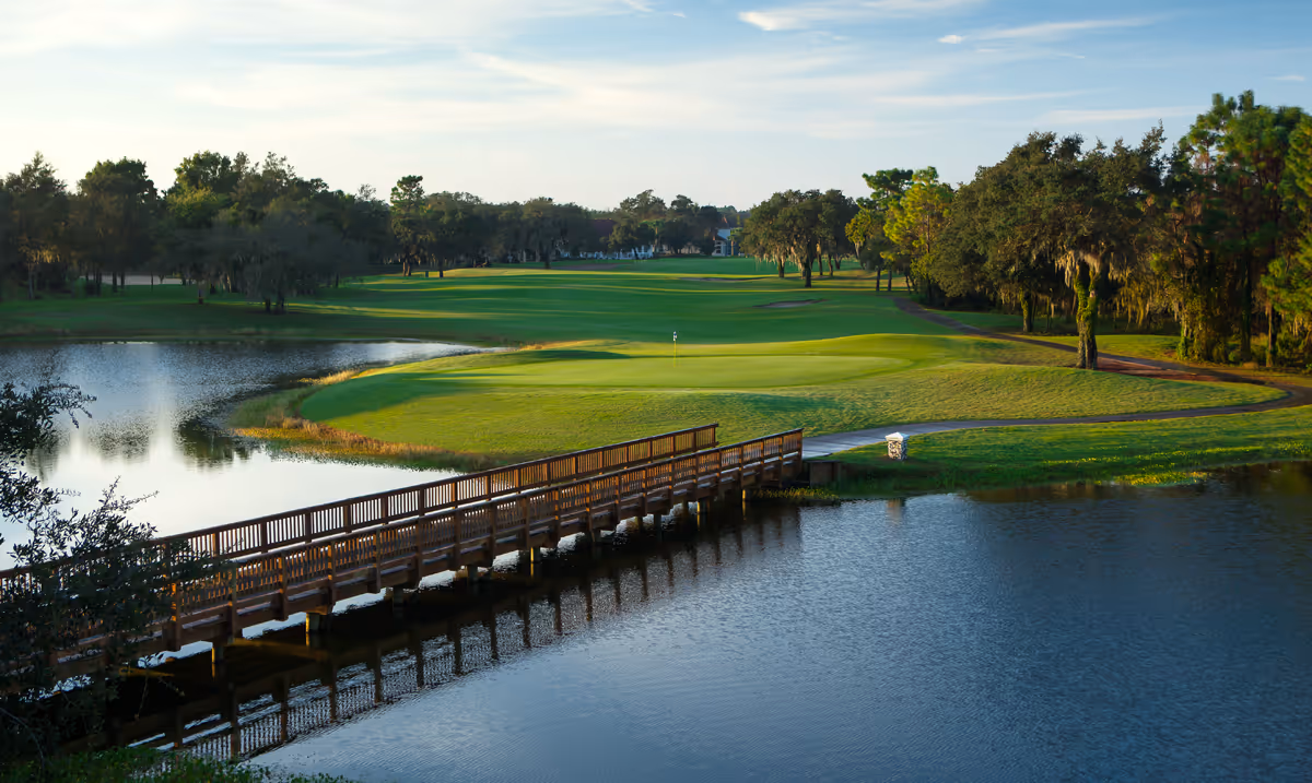 A scenic view of a golf course with a wooden bridge crossing over a water hazard. The green is well-maintained with a flagstick visible, surrounded by trees and a clear sky.