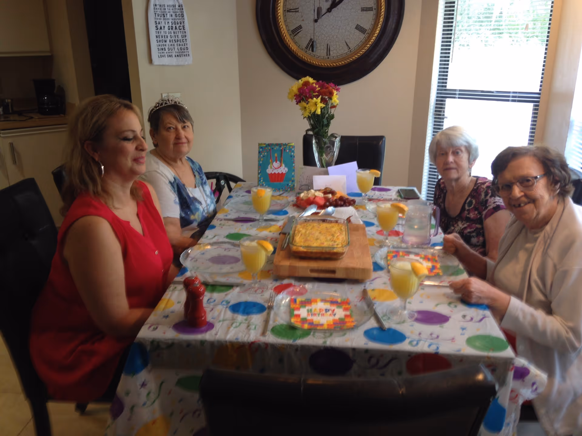 Four women sitting around a dining table decorated with a colorful tablecloth and birthday-themed plates. The table has glasses of orange juice with lemon slices, a casserole dish, a plate of fruit and cheese, and a vase with flowers. A large wall clock and a window with blinds are visible in the background.