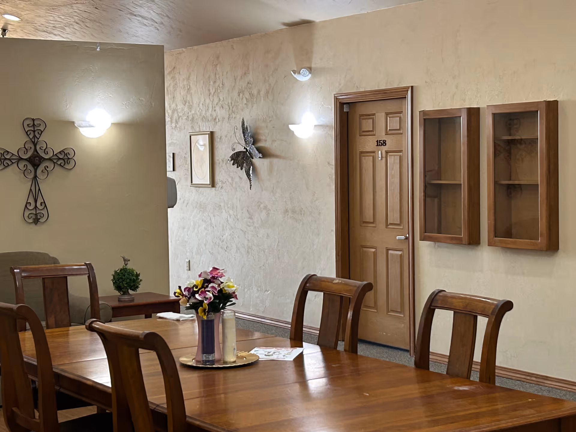Interior view of a senior living facility dining area with a wooden table and chairs. A vase with colorful flowers and a candle are placed on the table. The walls are textured and decorated with a metal cross, framed artwork, and a butterfly wall ornament. A wooden door labeled 158 and two glass-fronted wooden display cabinets are visible on the right wall.