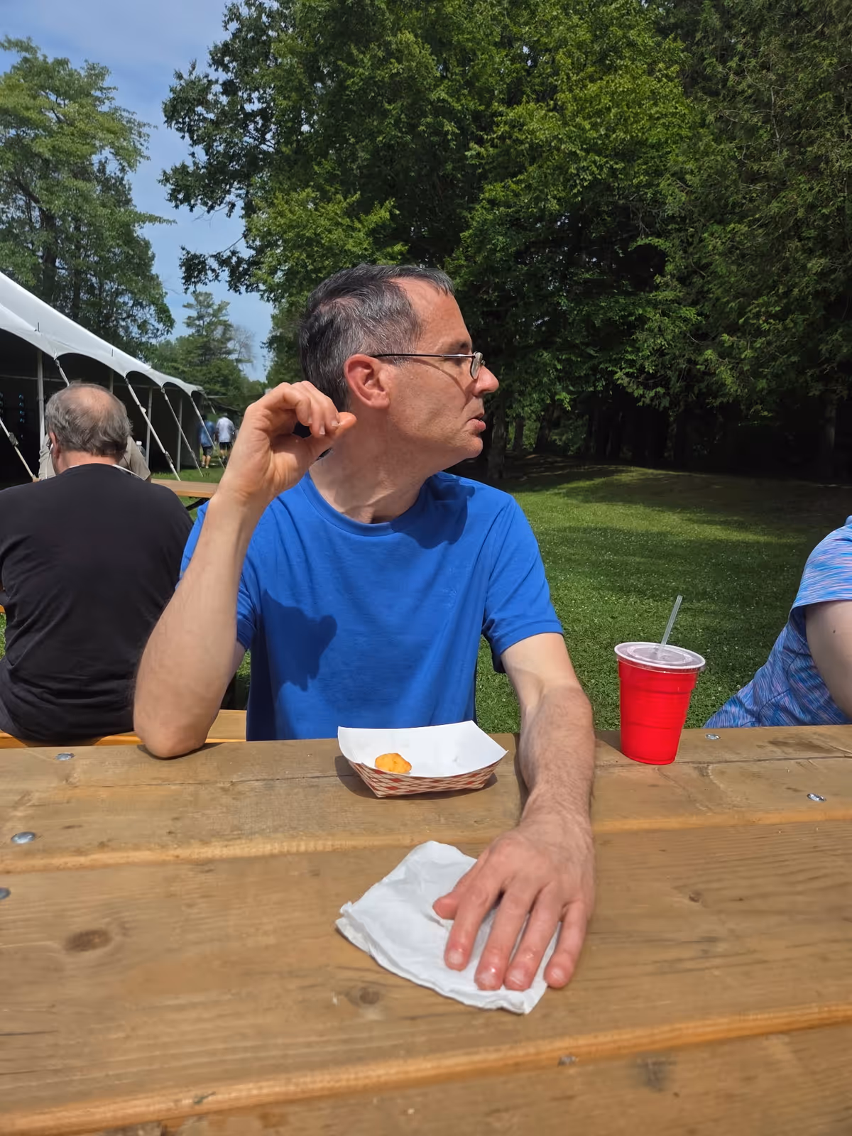 A man wearing glasses and a blue shirt sits at a wooden picnic table outdoors with a small food tray and a red cup with a straw in front of him. Behind him, there are trees, grass, and a white tent with other people sitting and walking nearby.