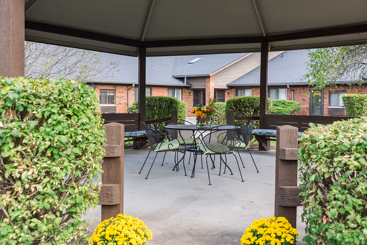 View of an outdoor gazebo with a round metal table and four chairs with cushions, surrounded by green bushes and yellow flowers, with a brick building in the background.