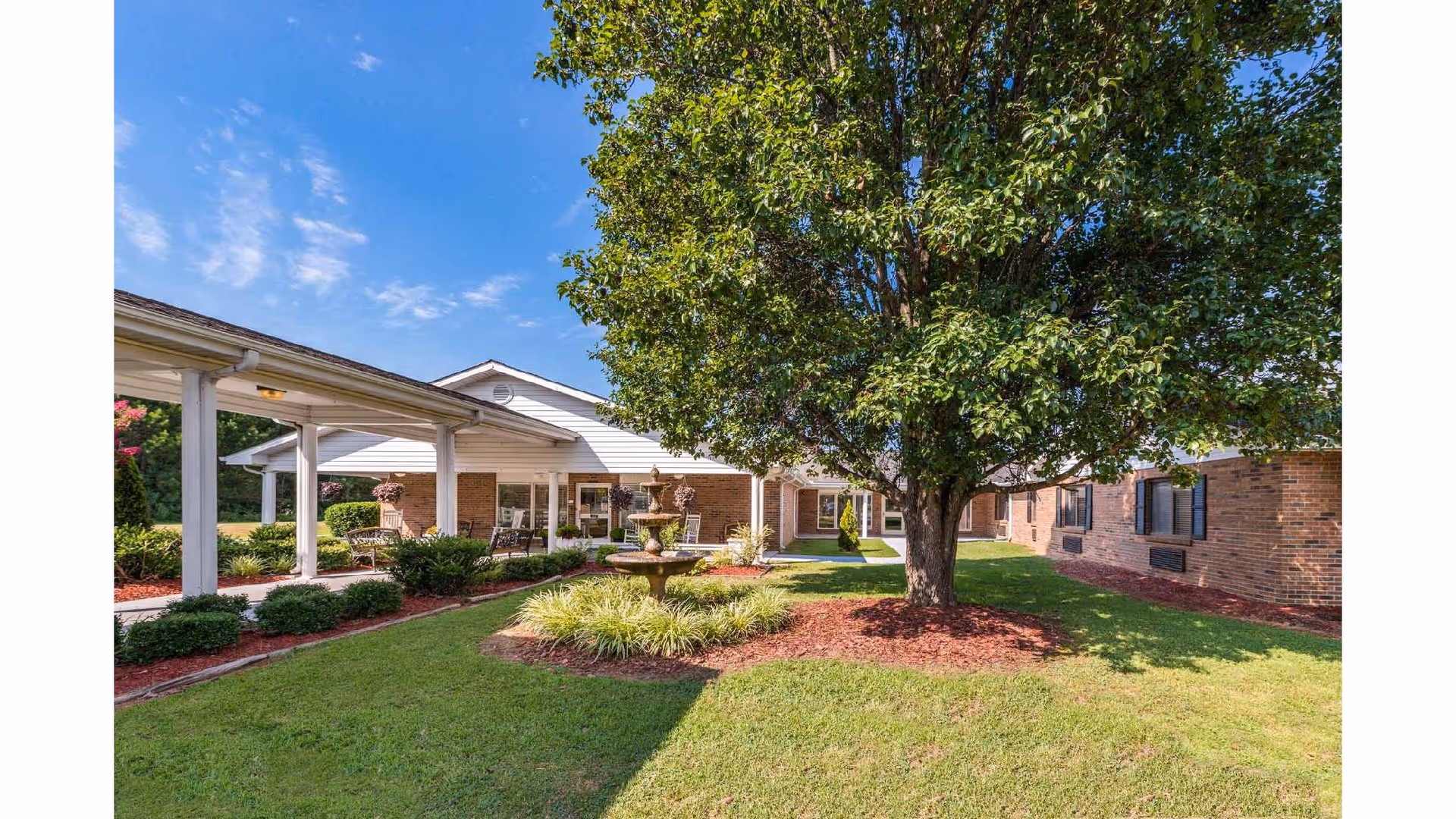 Outdoor courtyard area of Clayton Rehabilitation and Healthcare Center featuring a large tree, green lawn, a multi-tiered water fountain, and a covered walkway with seating. The building exterior is brick with white trim under a clear blue sky.