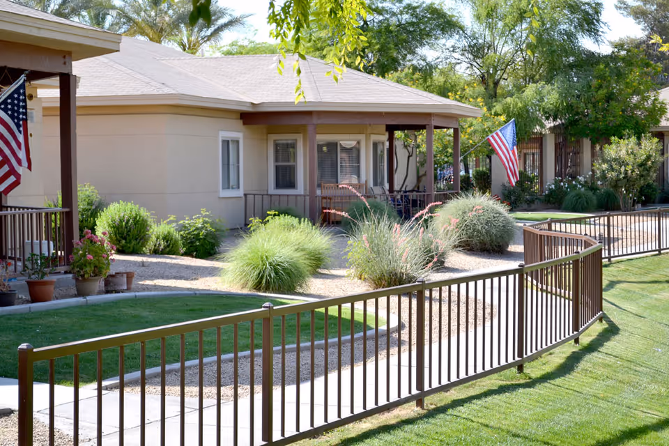 Front view of a single-story senior living cottage with a porch, landscaped courtyard, American flags and a curved metal railing.