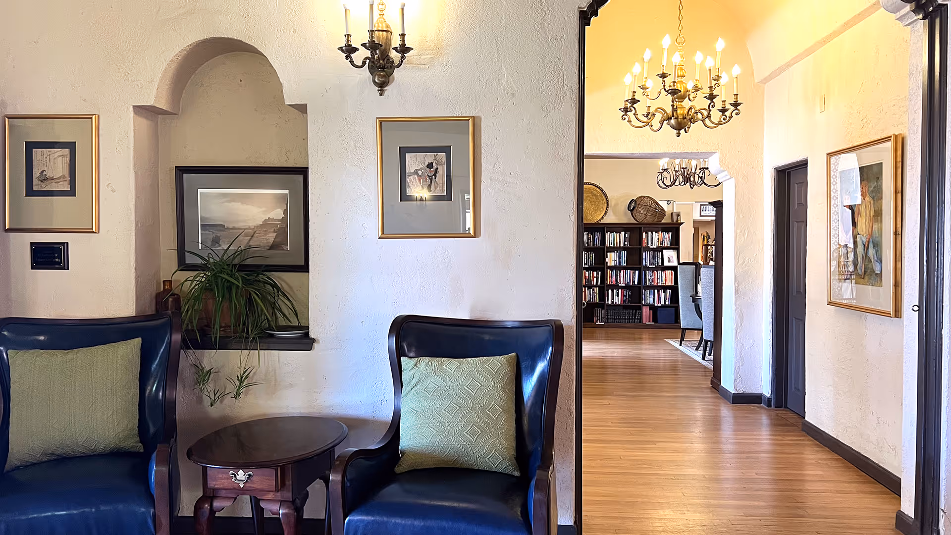 A cozy interior hallway with two dark blue armchairs with green cushions on either side of a small wooden table. The walls are textured and painted white, decorated with framed artwork. A small plant sits on a ledge in an arched wall niche. The hallway leads to a room with bookshelves and a dining area with chandeliers hanging from the ceiling.