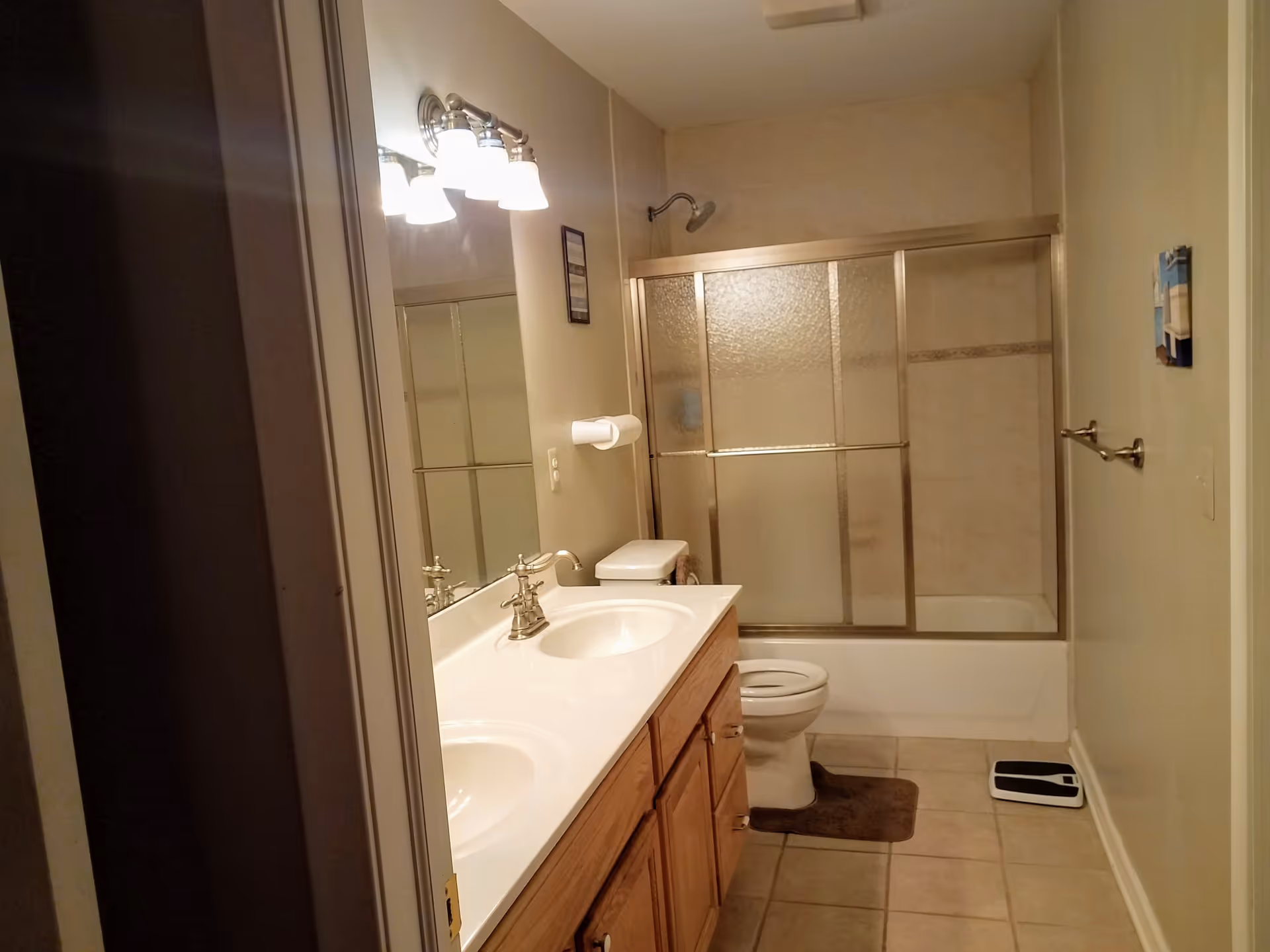 A bathroom with a double sink vanity with wooden cabinets, a large mirror above the sinks, a toilet, and a bathtub with sliding glass doors. The floor is tiled, and there is a brown bath mat in front of the toilet. A scale is placed on the floor near the wall, and a towel rack is mounted on the right wall.