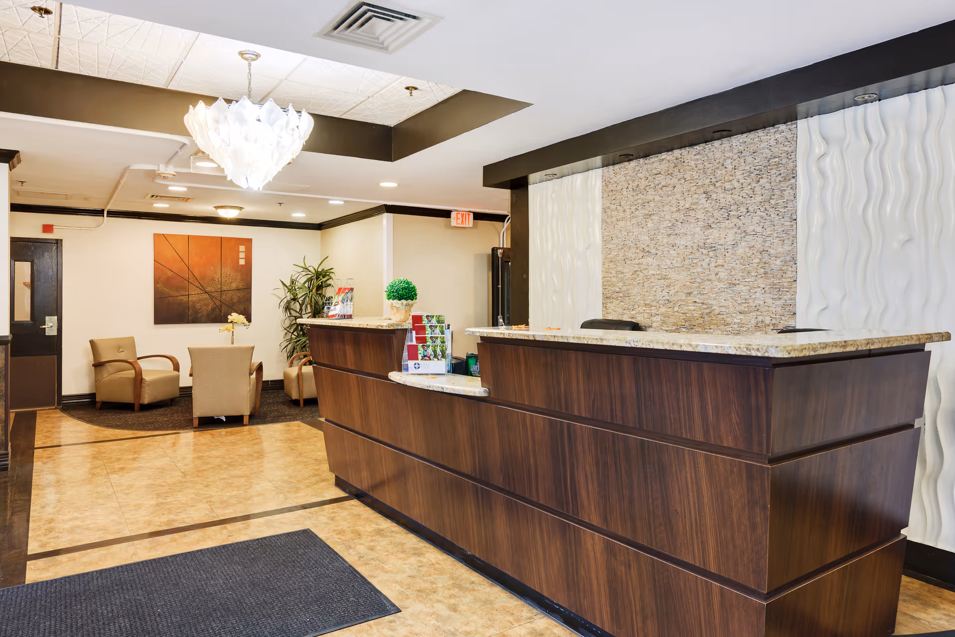Reception area of a senior living facility with a large wooden front desk, a modern chandelier hanging from the ceiling, and a seating area with beige chairs and a small table. The walls feature decorative panels and artwork, and there are informational brochures on the desk.