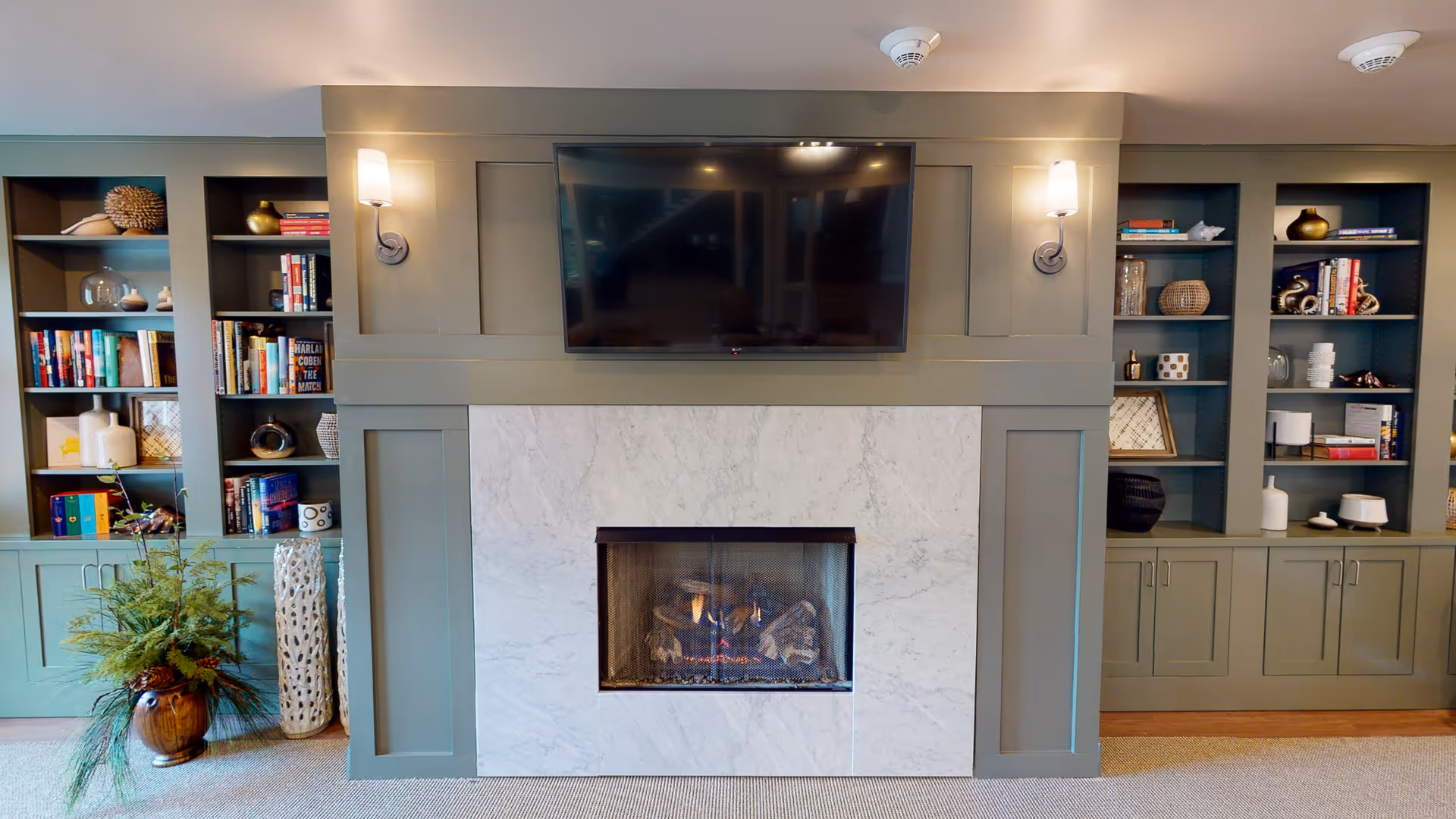 A cozy interior living room area featuring a modern fireplace with a marble surround and a flat-screen TV mounted above it. On either side of the fireplace are built-in shelves filled with books, decorative items, and vases. Two wall sconces provide warm lighting above the fireplace. A large potted plant is placed on the left side of the fireplace.