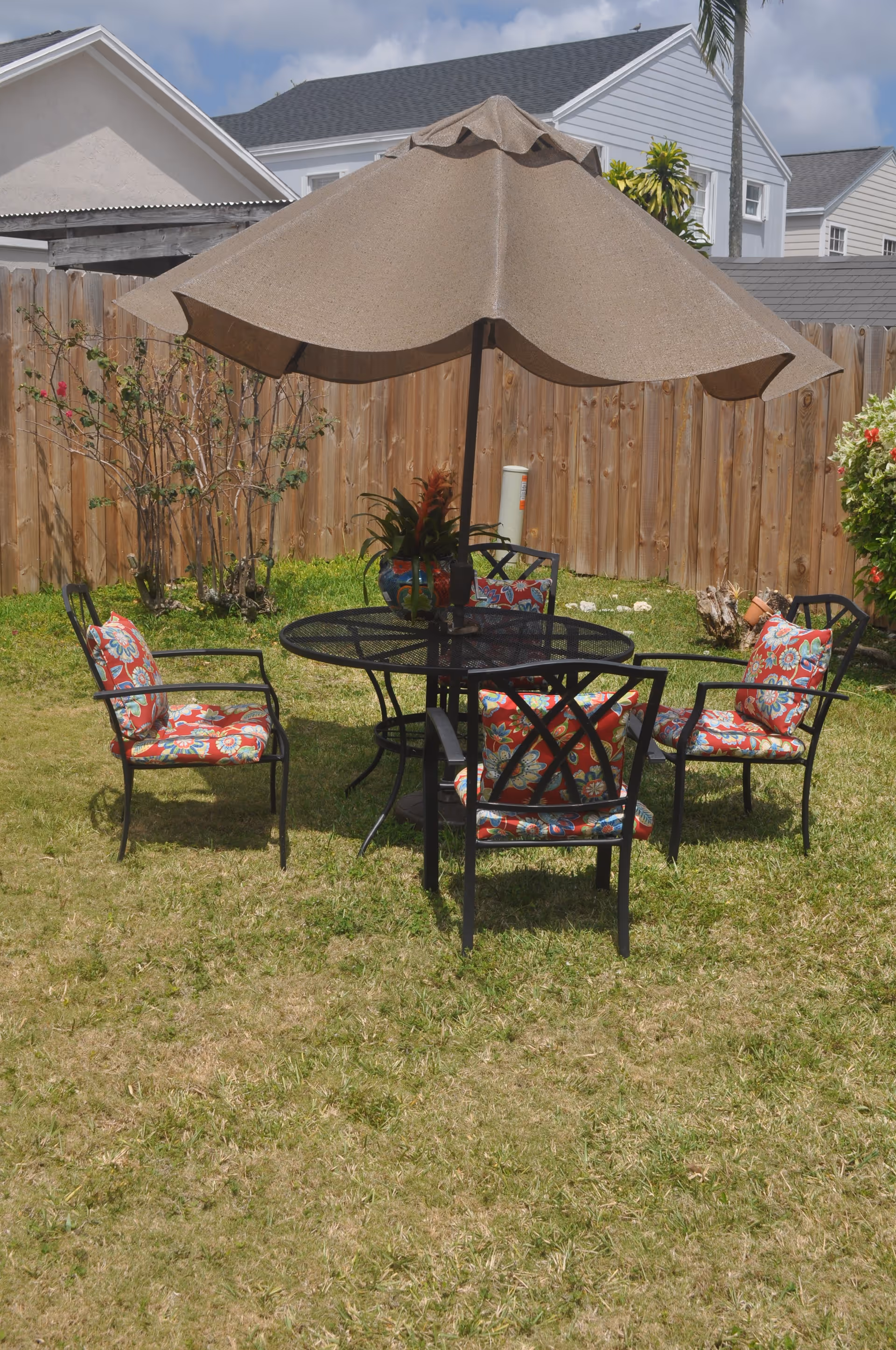 Backyard patio set with a round metal table, large umbrella and four floral-cushioned chairs on a grassy lawn in front of a wooden fence.