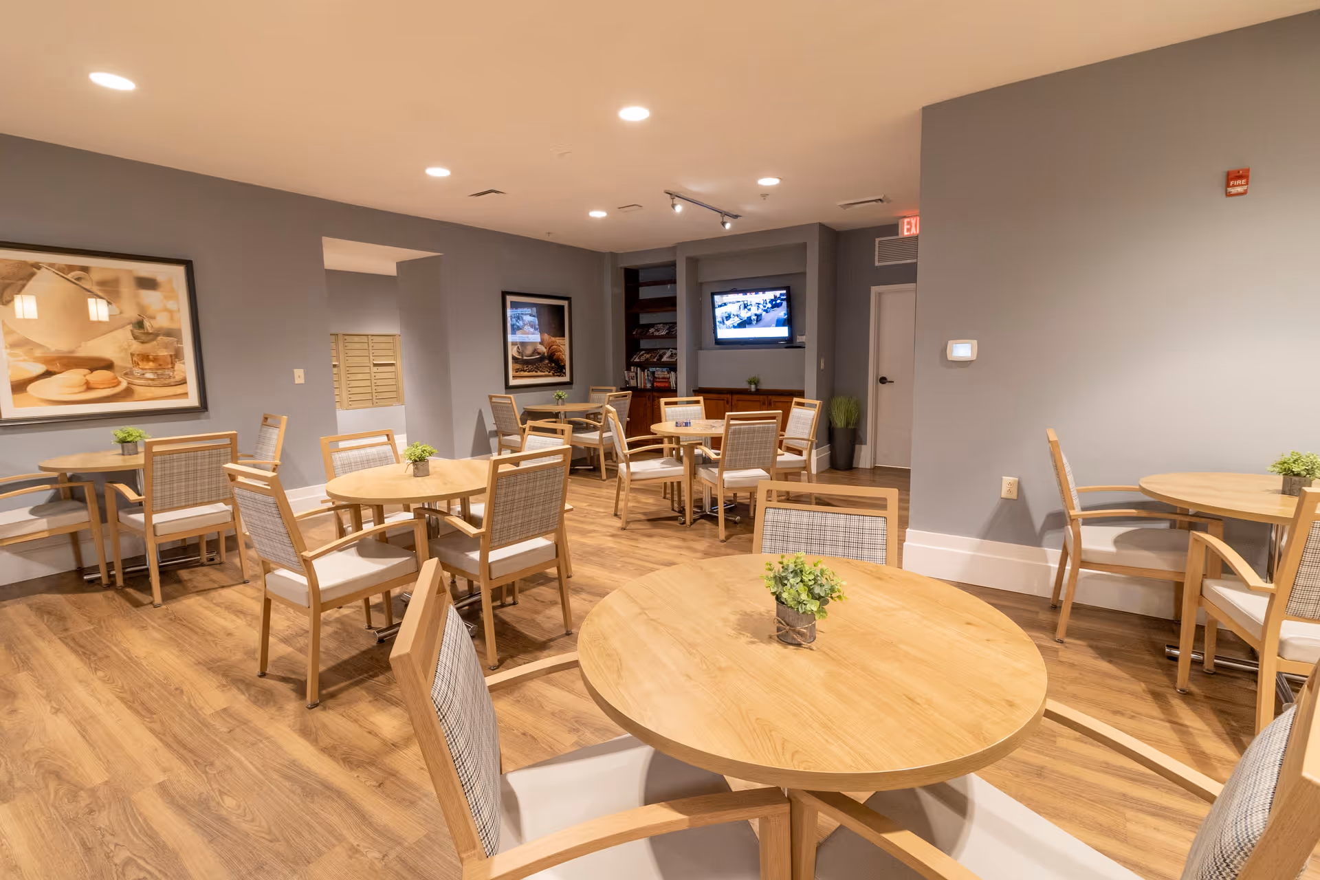 A dining room with several round wooden tables and chairs with light-colored cushions. The room has wood flooring, gray walls, framed pictures on the walls, and a television mounted on the far wall. Small green plants are placed on each table.