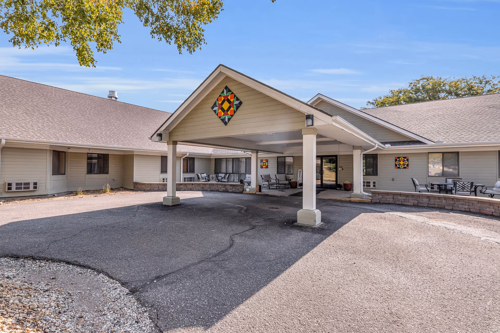 Exterior view of Cambridge Place senior living facility showing a covered entrance with seating areas on both sides, beige siding, and decorative colorful quilt-like patterns on the walls under a clear blue sky.