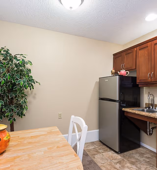 Small kitchen area with a wooden table and white chair in the foreground, a potted plant against a beige wall, a stainless steel refrigerator, wooden cabinets, and a countertop with a sink.