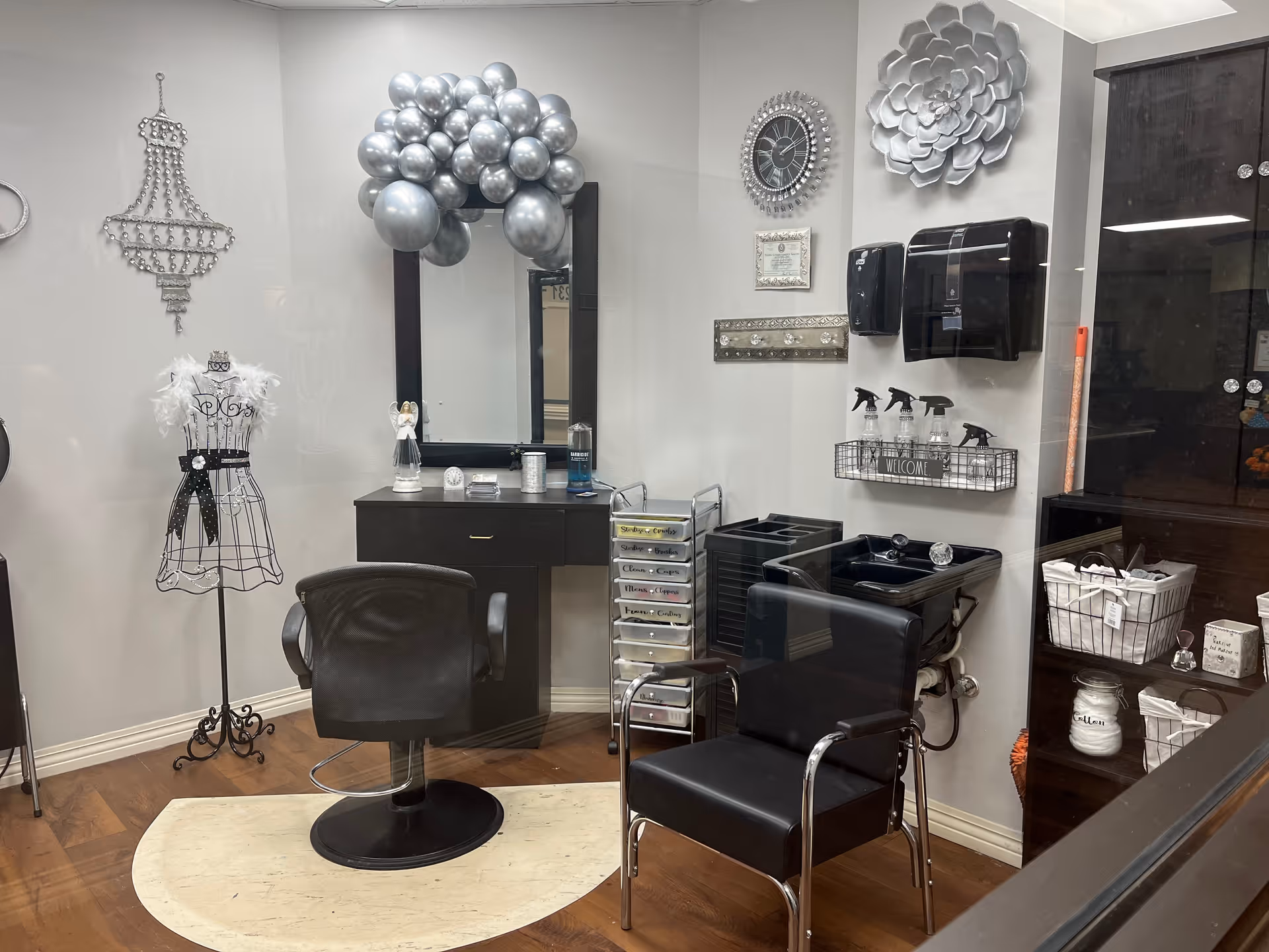 Interior view of a salon area with two black chairs, one in front of a vanity with a mirror decorated with silver balloons, and the other next to a black hair washing sink. The walls have decorative items including a clock, a large flower wall art, and a wire mannequin with a feather boa. Shelves and baskets with salon supplies are visible.