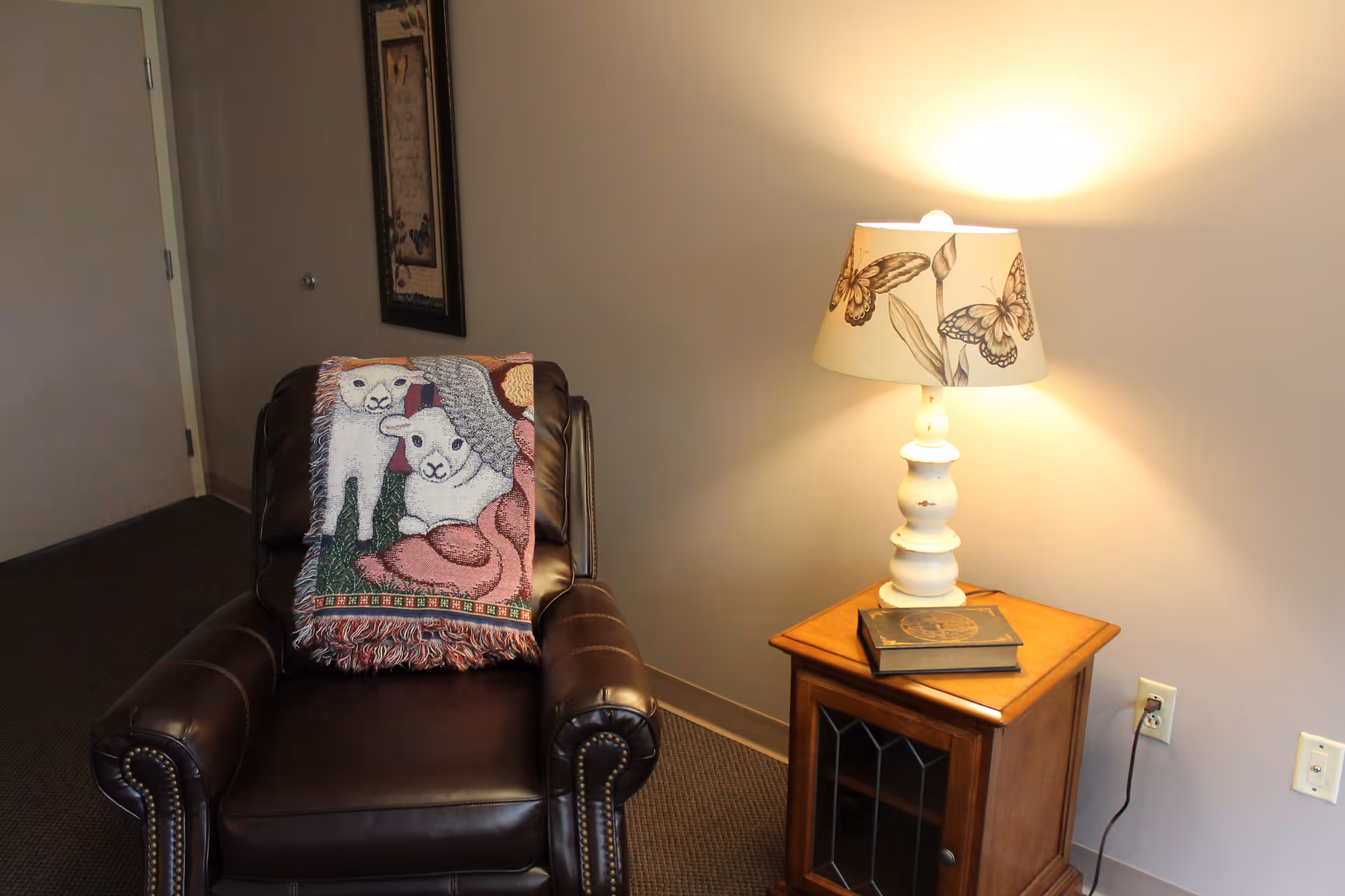 Leather recliner with a sheep-themed throw next to a wooden side table topped by a lamp and book in an interior room.