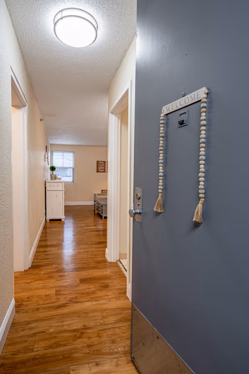 View looking down a hallway with wooden flooring and beige walls. On the right side, a gray door is partially open with a decorative 'WELCOME' sign hanging on it. At the end of the hallway, there is a white cabinet with a small plant on top and a window above it. A coffee table and wall decor are visible in the room beyond the hallway.