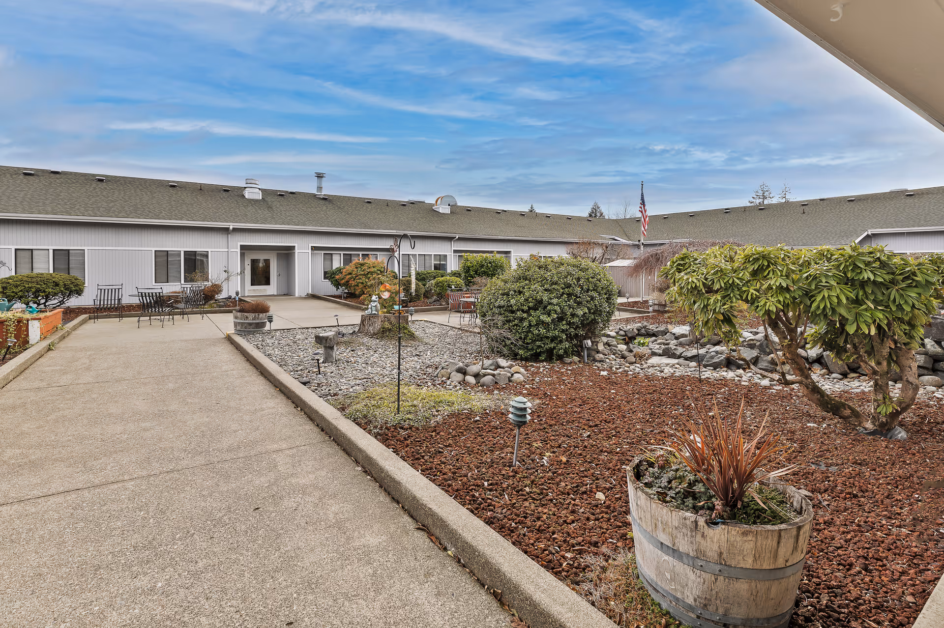 Outdoor courtyard area of a senior living facility with a paved walkway, landscaped garden beds with shrubs and small trees, outdoor seating with tables and chairs, and a building with multiple windows and doors surrounding the courtyard under a partly cloudy sky.