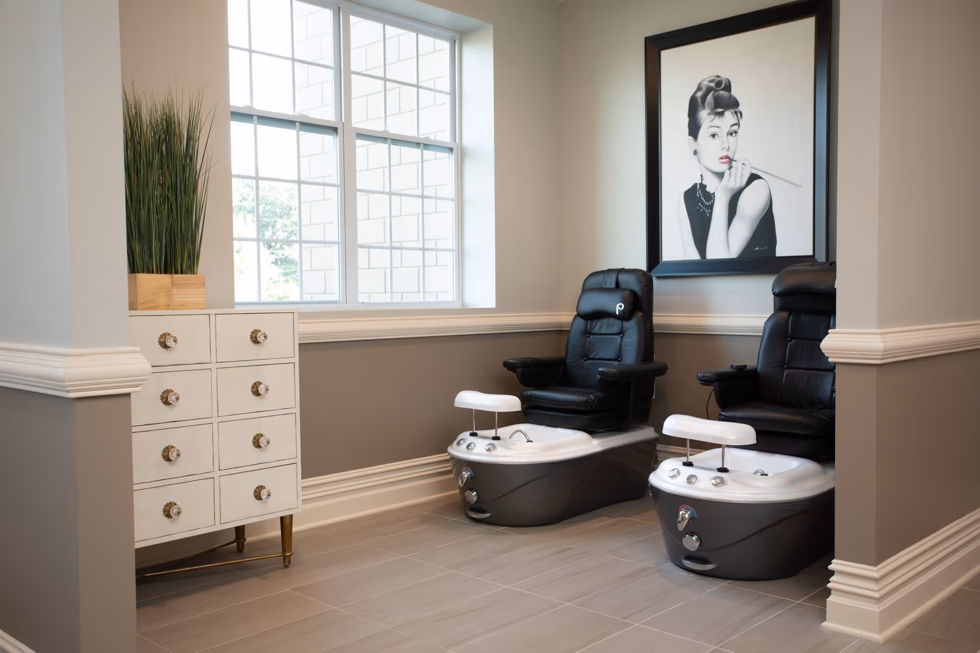 A modern pedicure station with two black leather chairs and white foot baths in a well-lit room. There is a white cabinet with drawers and a tall green plant on top next to a large window. A framed black and white portrait of a woman smoking a cigarette is hung on the wall.