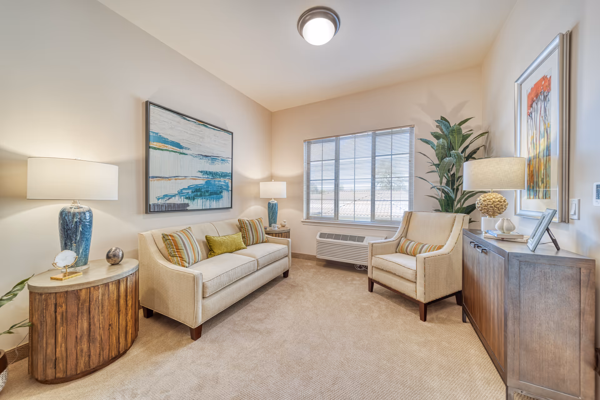A cozy living room in a senior living facility featuring a beige sofa with striped and green cushions, a matching armchair, two wooden side tables with lamps, a large window with blinds, a potted plant, and framed artwork on the walls.