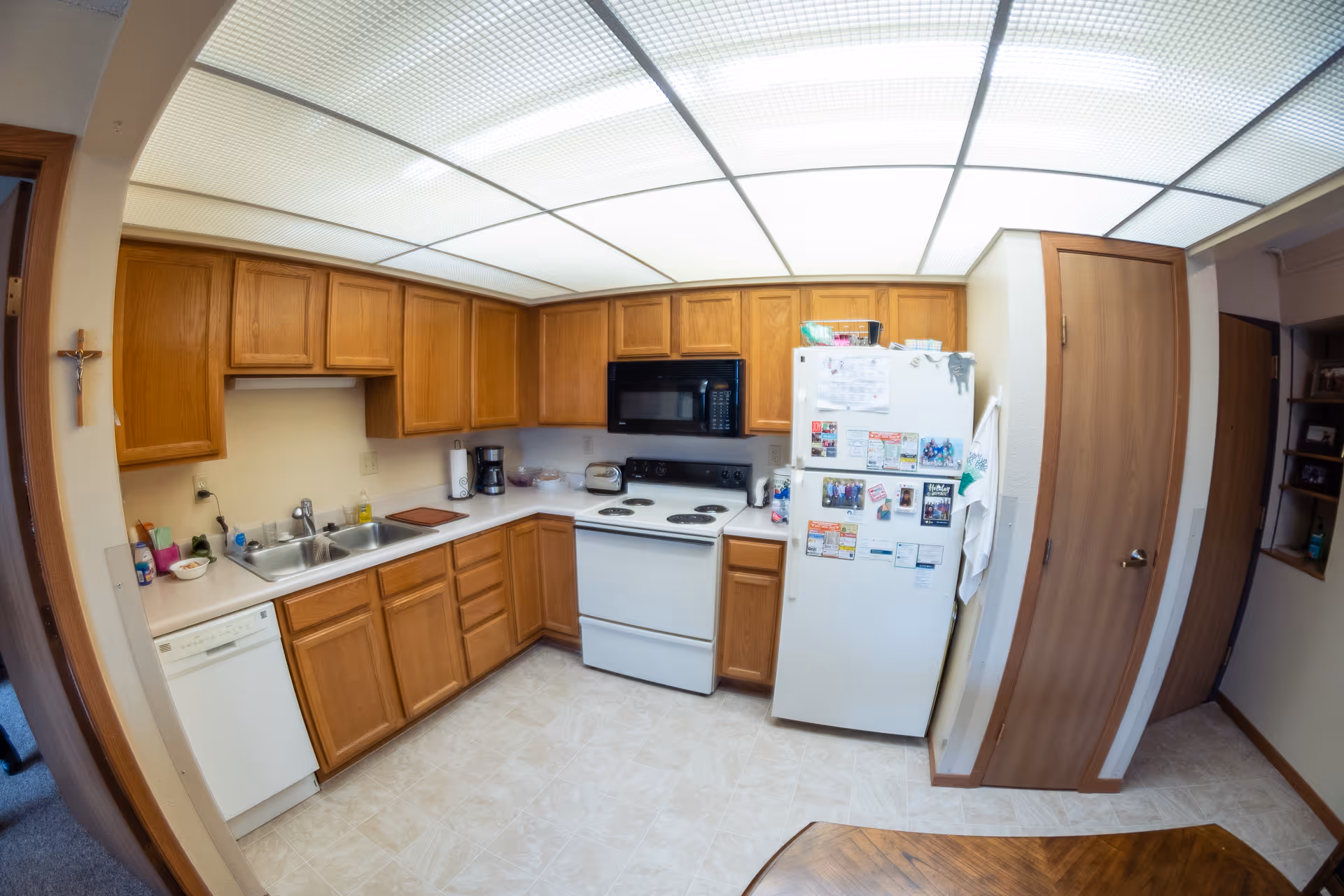 A well-lit kitchen with wooden cabinets, a white refrigerator covered with magnets and papers, a white electric stove with a black microwave above it, a double sink, a dishwasher, and various kitchen items on the countertops. The ceiling has large fluorescent lights, and there is a wooden door and a small window with shelves on the right side.