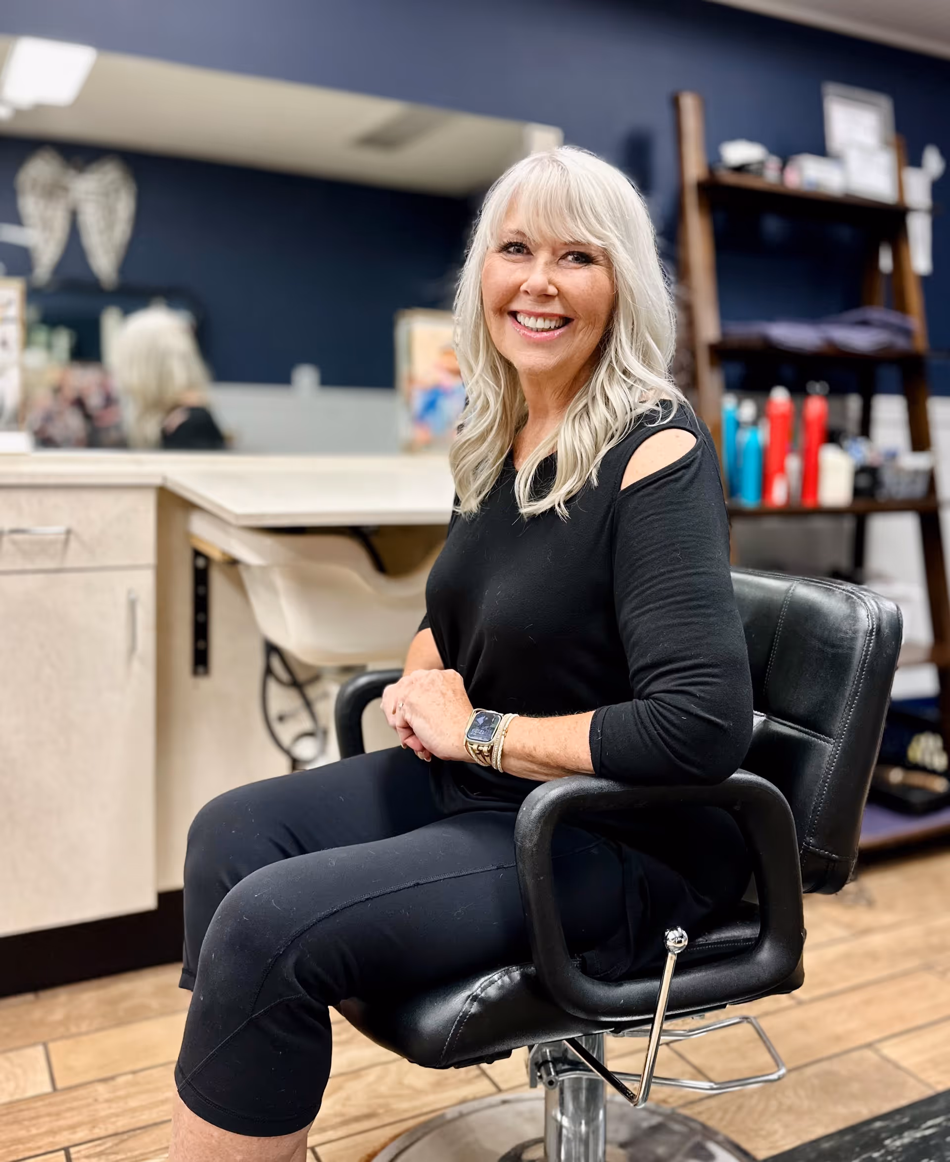 A smiling older woman with long white hair sitting in a black salon chair inside a hair salon. The background shows a mirror, a counter, and shelves with hair products.