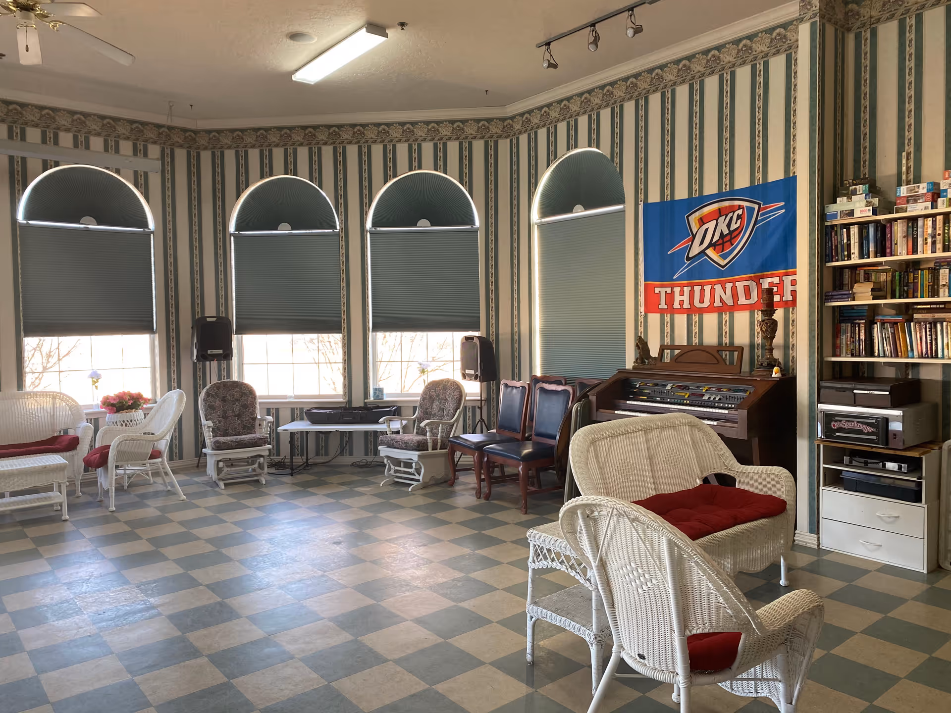 Sunlit common room with wicker seating, upholstered chairs, a bookshelf and an organ beneath arched windows.