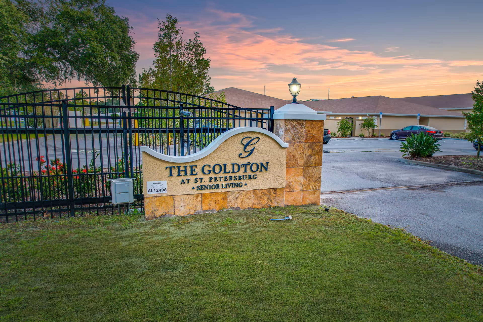 Entrance sign for The Goldton at St. Petersburg senior living facility with a gated fence, a lamp post on the sign, and a parking lot with cars in the background during sunset.