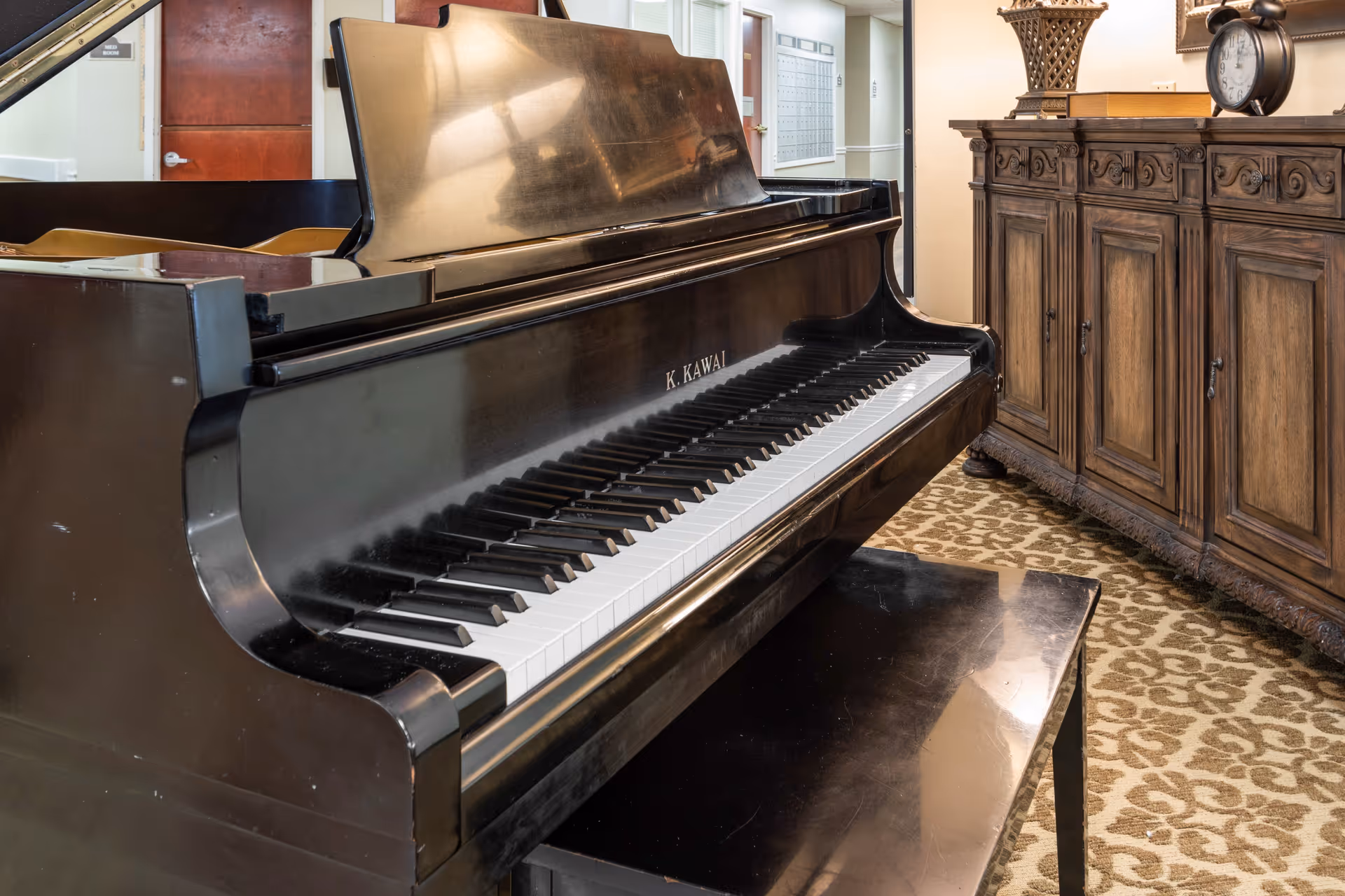 Close-up view of a black grand piano with the brand name K. Kawai visible on the side, placed on a patterned carpet in a room with a wooden cabinet and an alarm clock on top.