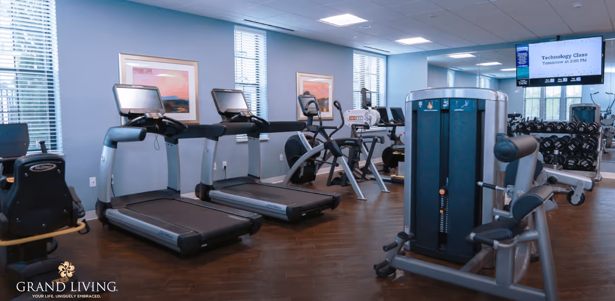 Interior view of a fitness center with treadmills, exercise bikes, weight machines, and dumbbells. The room has large windows with blinds, framed artwork on the walls, and a TV screen displaying information about a technology class.
