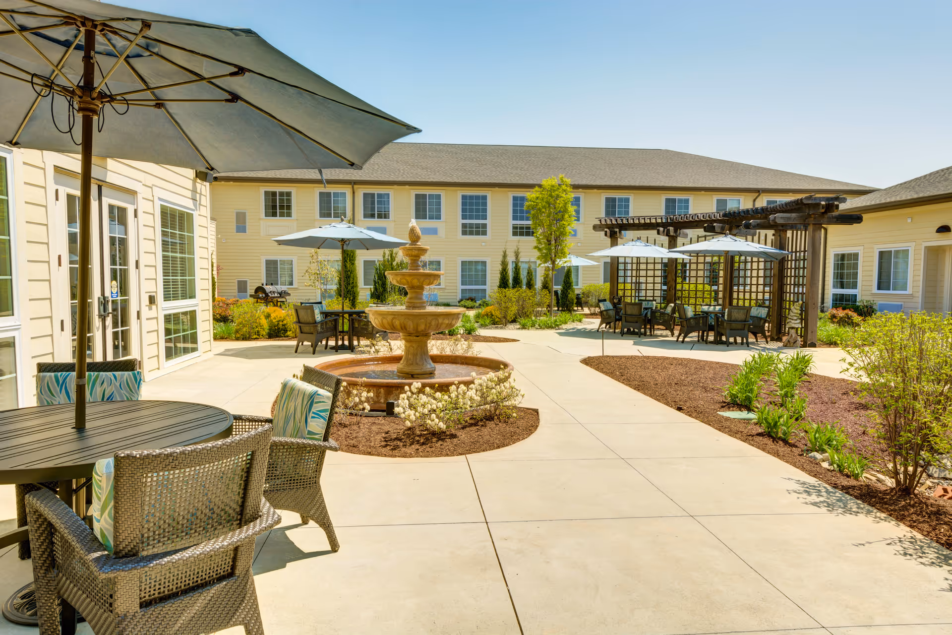 Outdoor courtyard area of a senior living facility with multiple seating areas featuring tables, chairs, and umbrellas. A three-tiered water fountain is centrally located, surrounded by landscaped plants and shrubs. The building with multiple windows surrounds the courtyard under a clear blue sky.